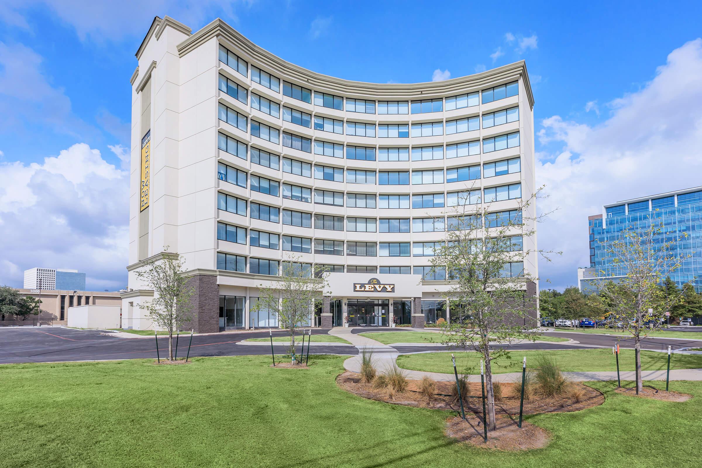 A modern multi-story building with a curved design featuring large windows. The facade has a sign reading "LEVY." In front of the building, there is a landscaped area with small trees and grass, set against a cloudy blue sky.
