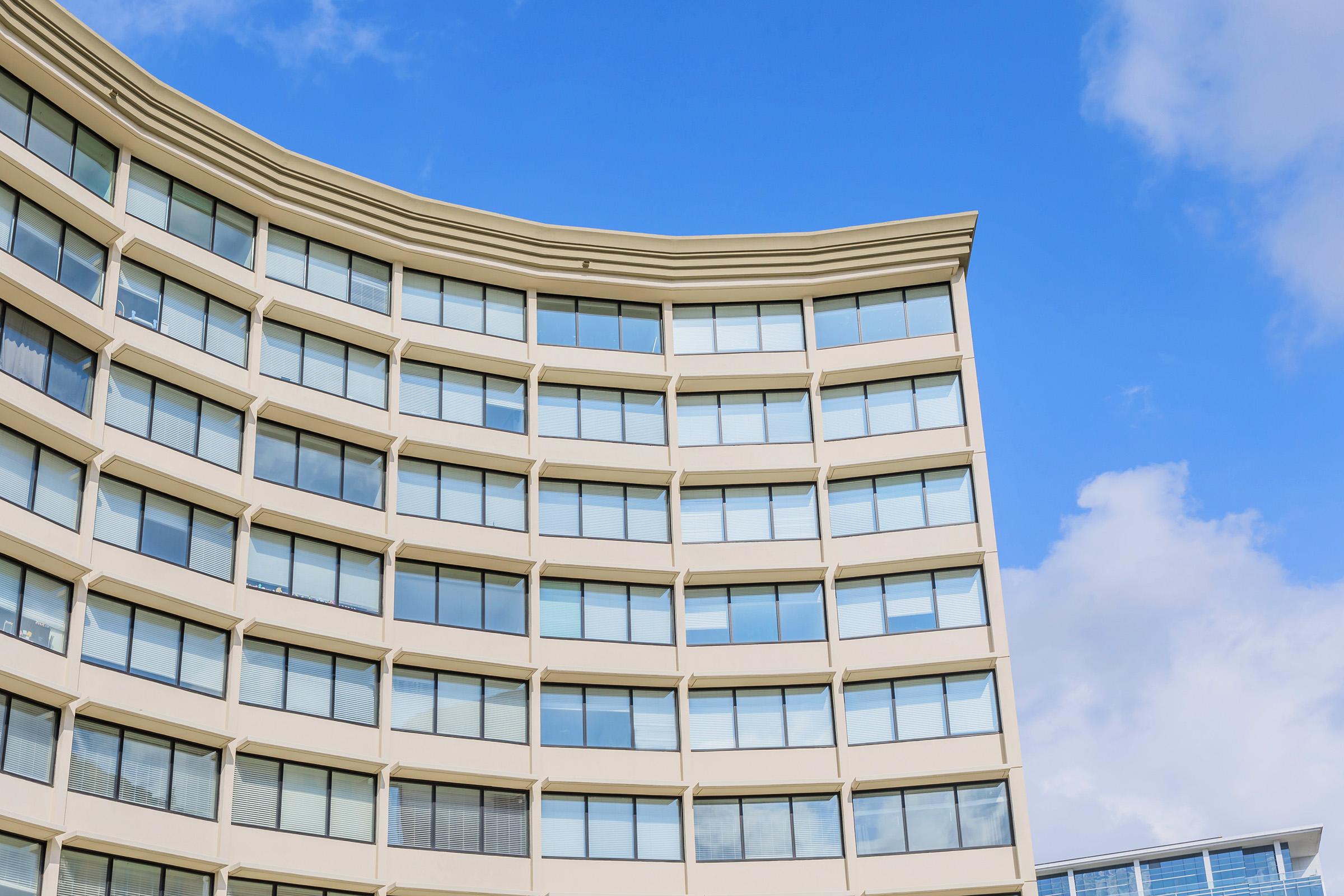 A modern building with a curved facade featuring large windows, set against a bright blue sky with a few fluffy clouds. The architectural design highlights the smooth lines and contemporary style of the structure.