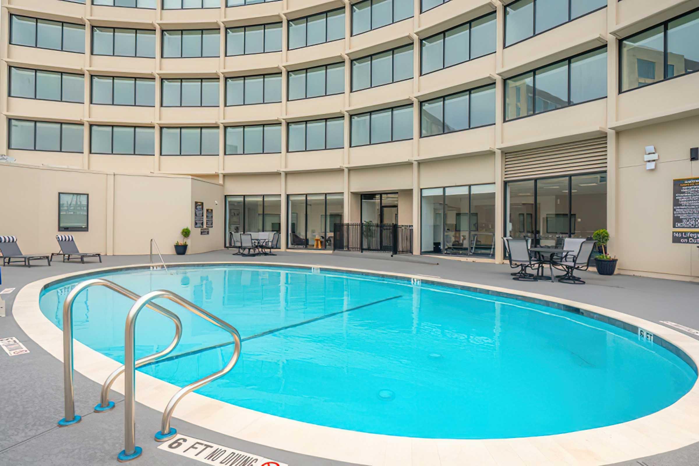 A modern hotel pool area featuring a round-shaped blue pool with a handrail. Surrounding the pool are patio chairs, and large windows are visible in the background, suggesting an indoor-outdoor experience. The ambiance is bright and inviting, perfect for relaxation.