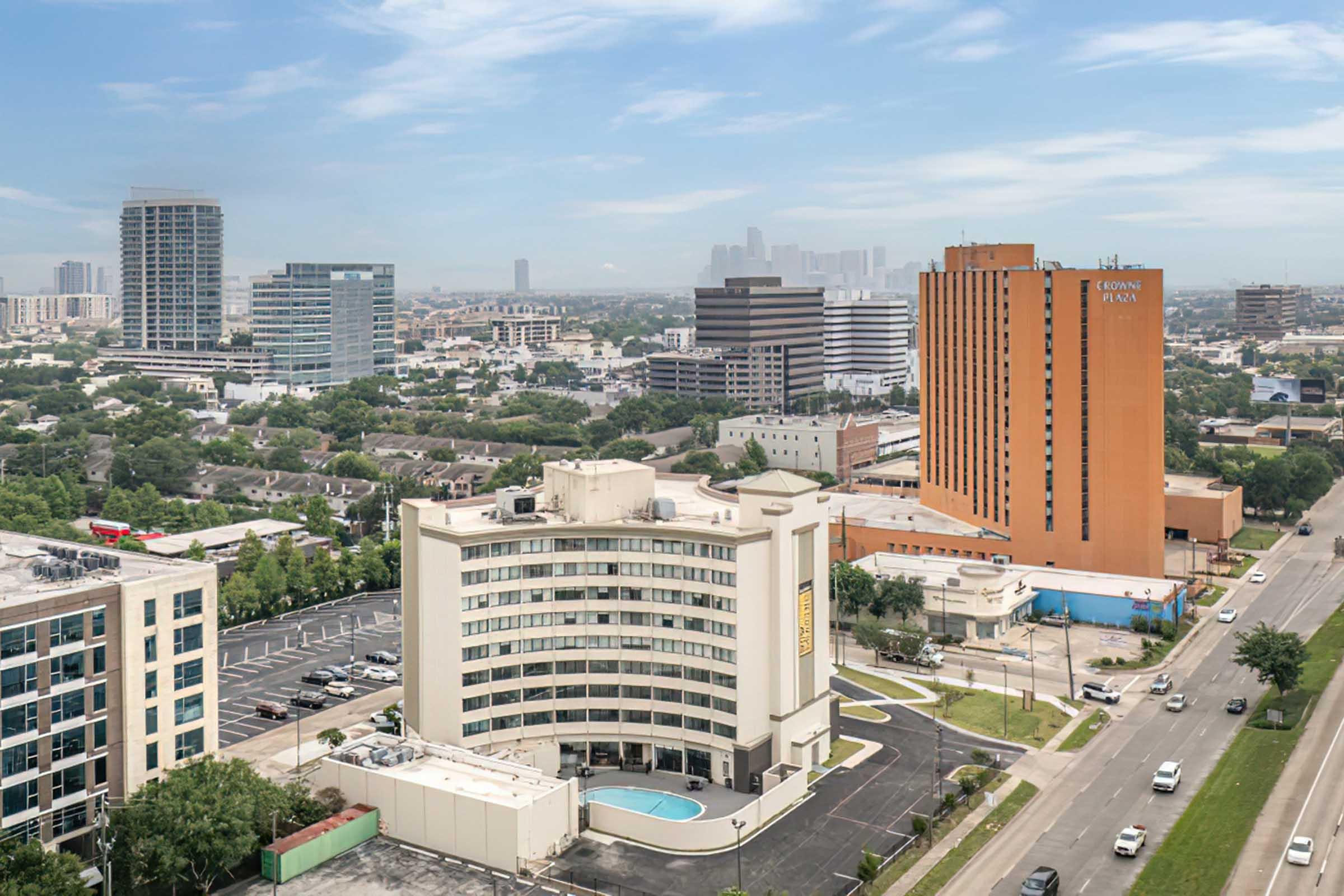 Aerial view of a city skyline featuring a mix of high-rise buildings and low-rise structures, with an orange hotel prominently displayed. Green trees and a parking lot are visible in the foreground, alongside a swimming pool. The background showcases more urban development under a partly cloudy sky.