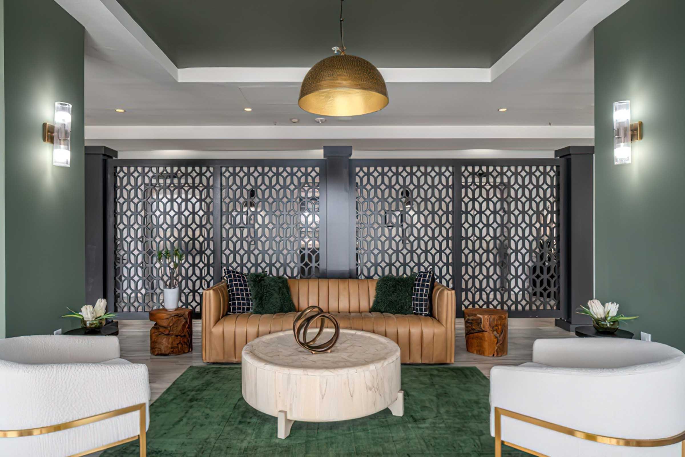 A stylish lounge area featuring a brown leather sofa with black and white patterned cushions, two white armchairs, and a round wooden coffee table. The space has green walls, decorative screens, and modern lighting fixtures, creating an inviting, contemporary atmosphere. A green area rug adds warmth to the design.