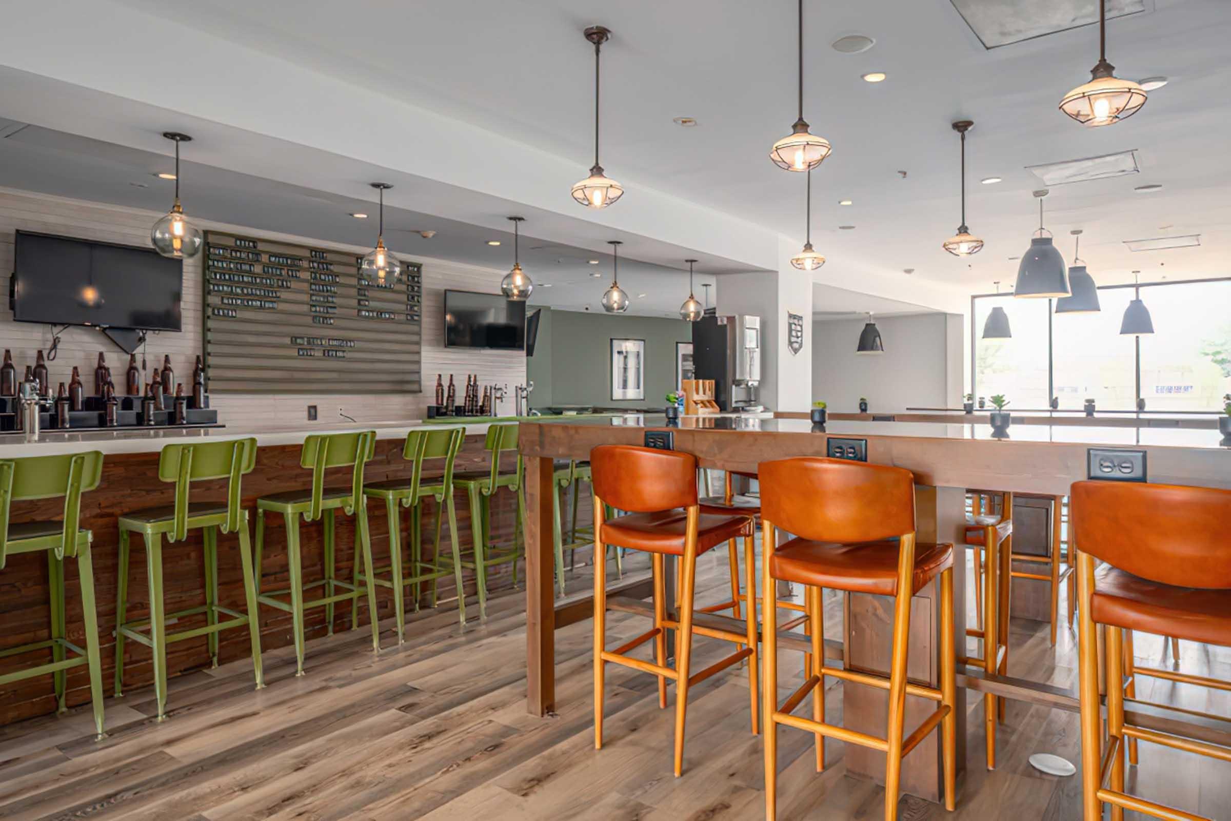 A modern, brightly lit bar area featuring high tables and stools in warm wood tones, with green and orange seating. Light fixtures hang from the ceiling, and walls have a mix of wood and neutral colors. A beer list is displayed on one wall, and the space has large windows allowing natural light in.