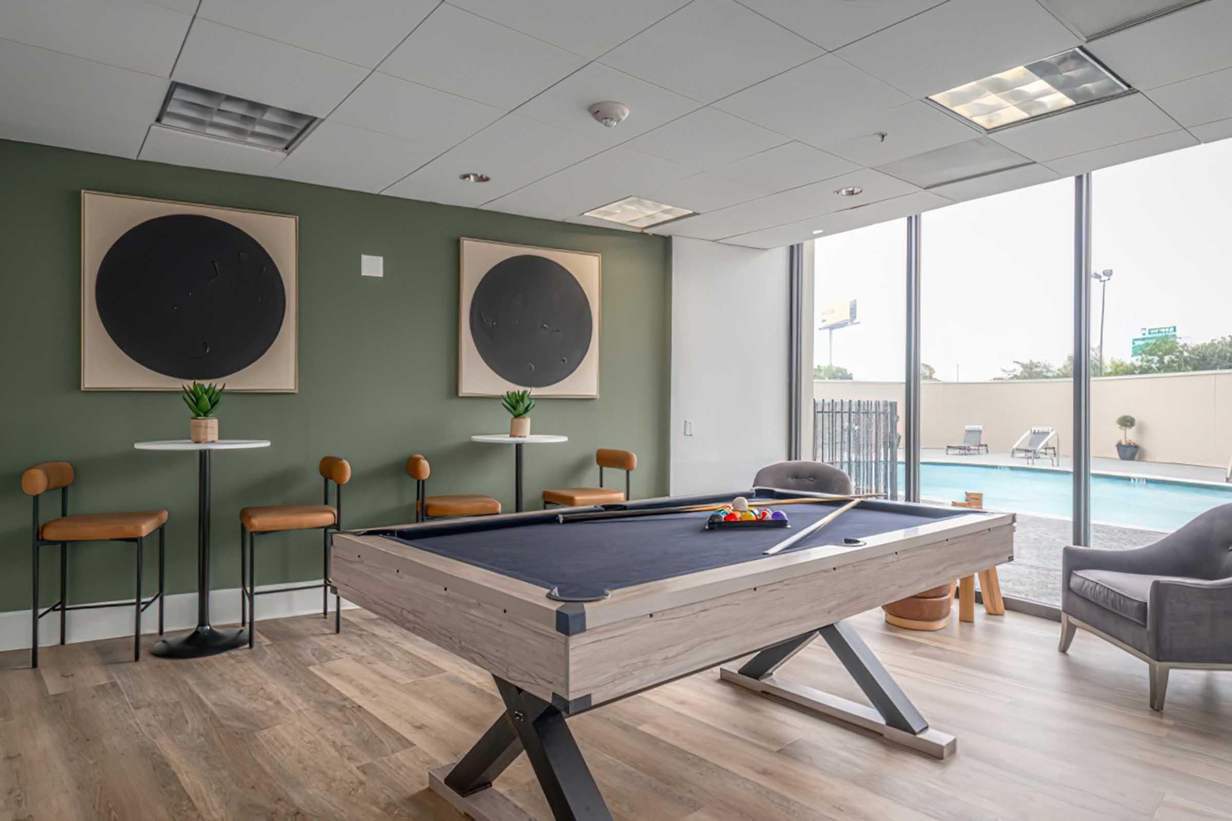 A modern recreational room featuring a pool table with a colorful set of balls, two small round tables with barstools, and a comfortable armchair. The walls are painted green, and there are two abstract black circle artworks. Large windows offer a view of an outdoor pool area.