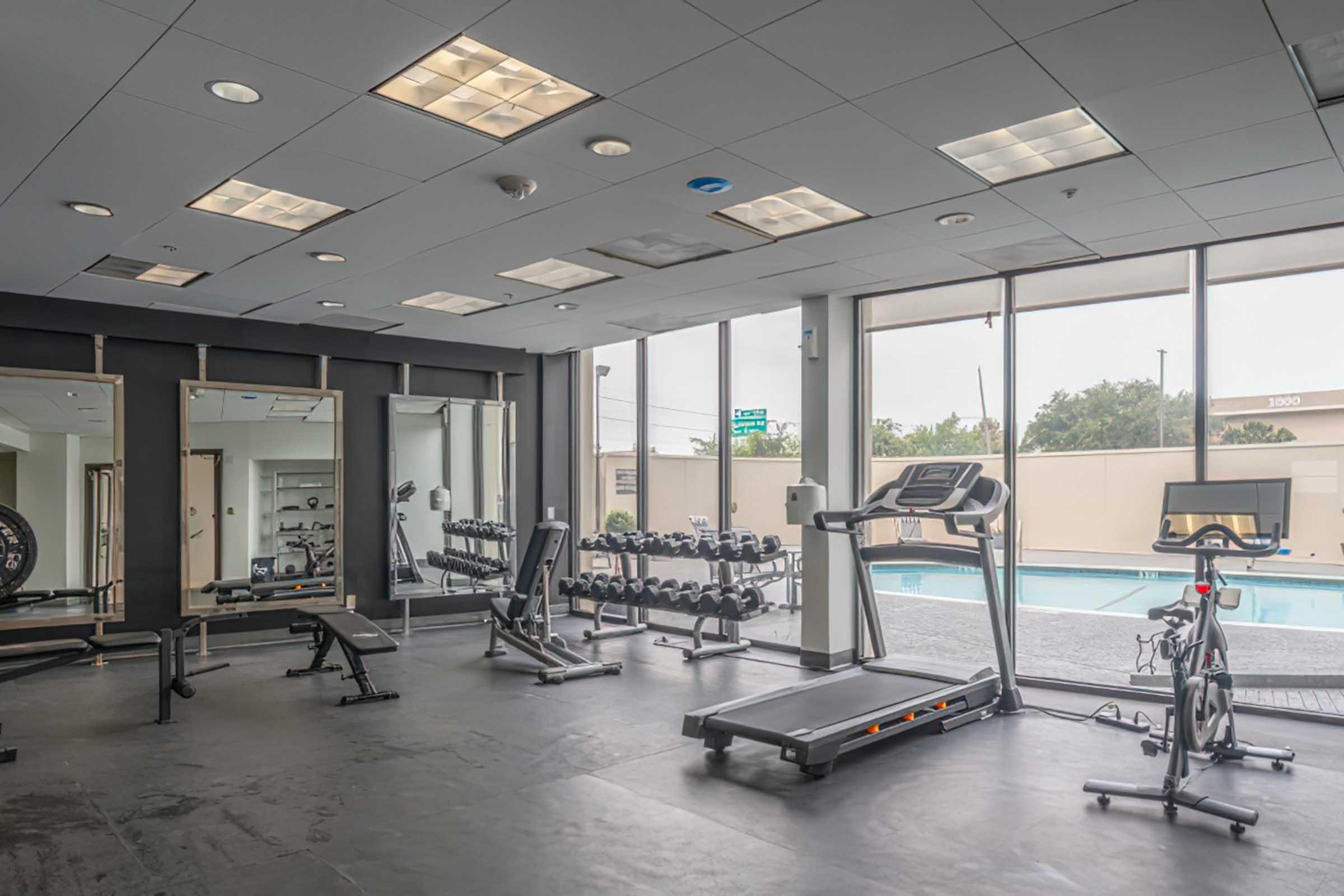 A modern gym interior featuring various exercise equipment. There are weight benches, dumbbells, a treadmill, and a stationary bike. Large windows let in natural light, providing a view of an outdoor swimming pool. Mirrors line one wall, enhancing the open feel of the space.