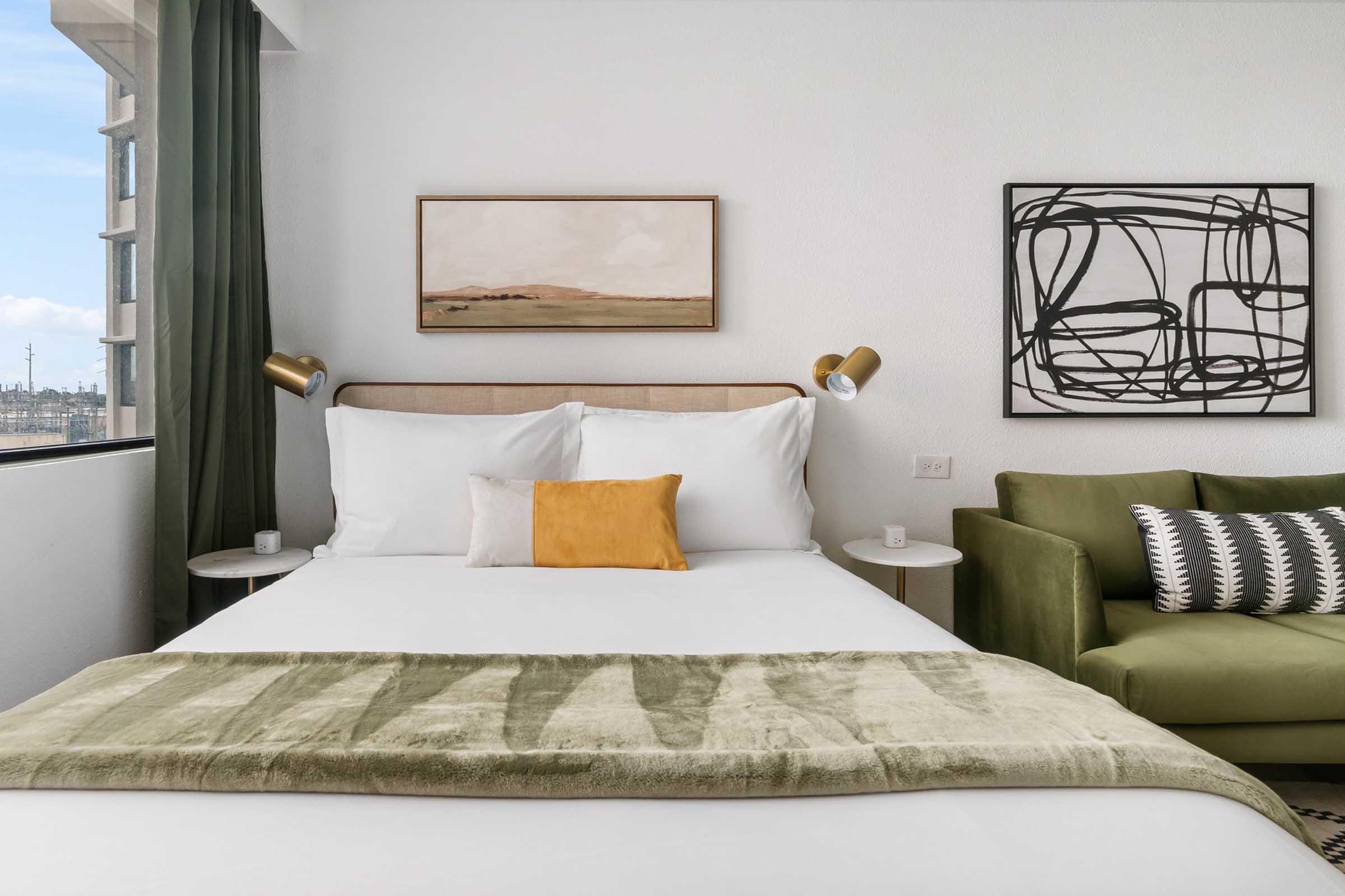 A cozy hotel room featuring a queen-sized bed with white linens and a green throw blanket. There are two bedside lamps, a small orange accent pillow, and abstract artwork on the walls. A green sofa with patterned cushions is positioned beside the bed, with large windows allowing natural light to illuminate the space.