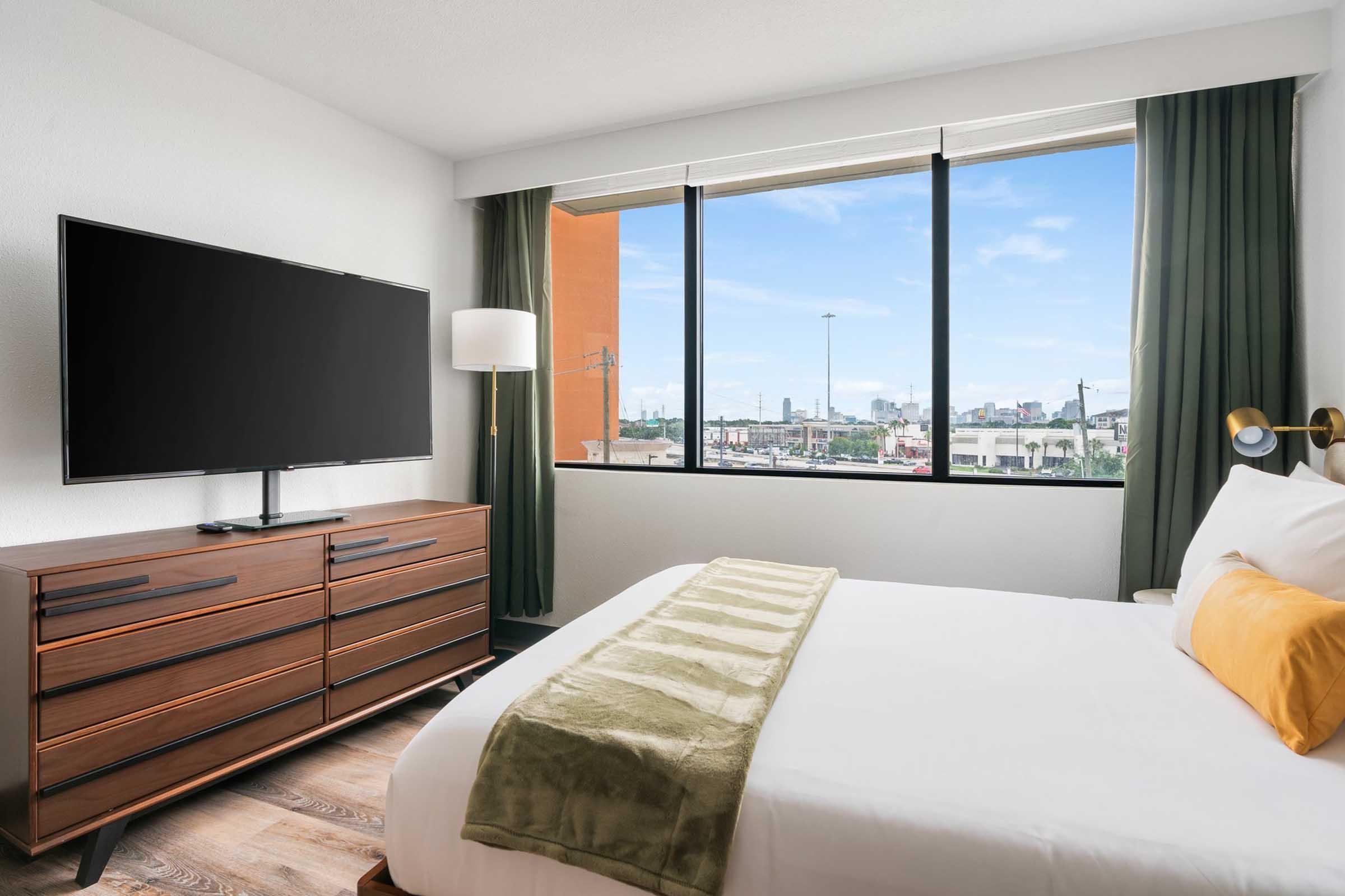 A modern hotel room featuring a comfortable bed with a green throw blanket, a stylish wooden dresser with a flat-screen TV on top, and large windows with green curtains allowing natural light and a view of the outside city.