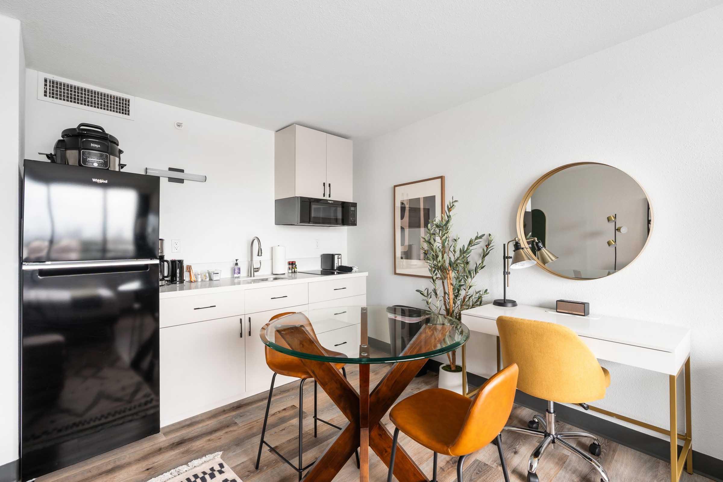 A modern studio apartment featuring a kitchenette with a black refrigerator, white cabinets, and a glass dining table surrounded by yellow chairs. There's a round mirror on the wall, a small desk with a lamp, and a decorative plant in the corner, all set in a bright and minimalist style.
