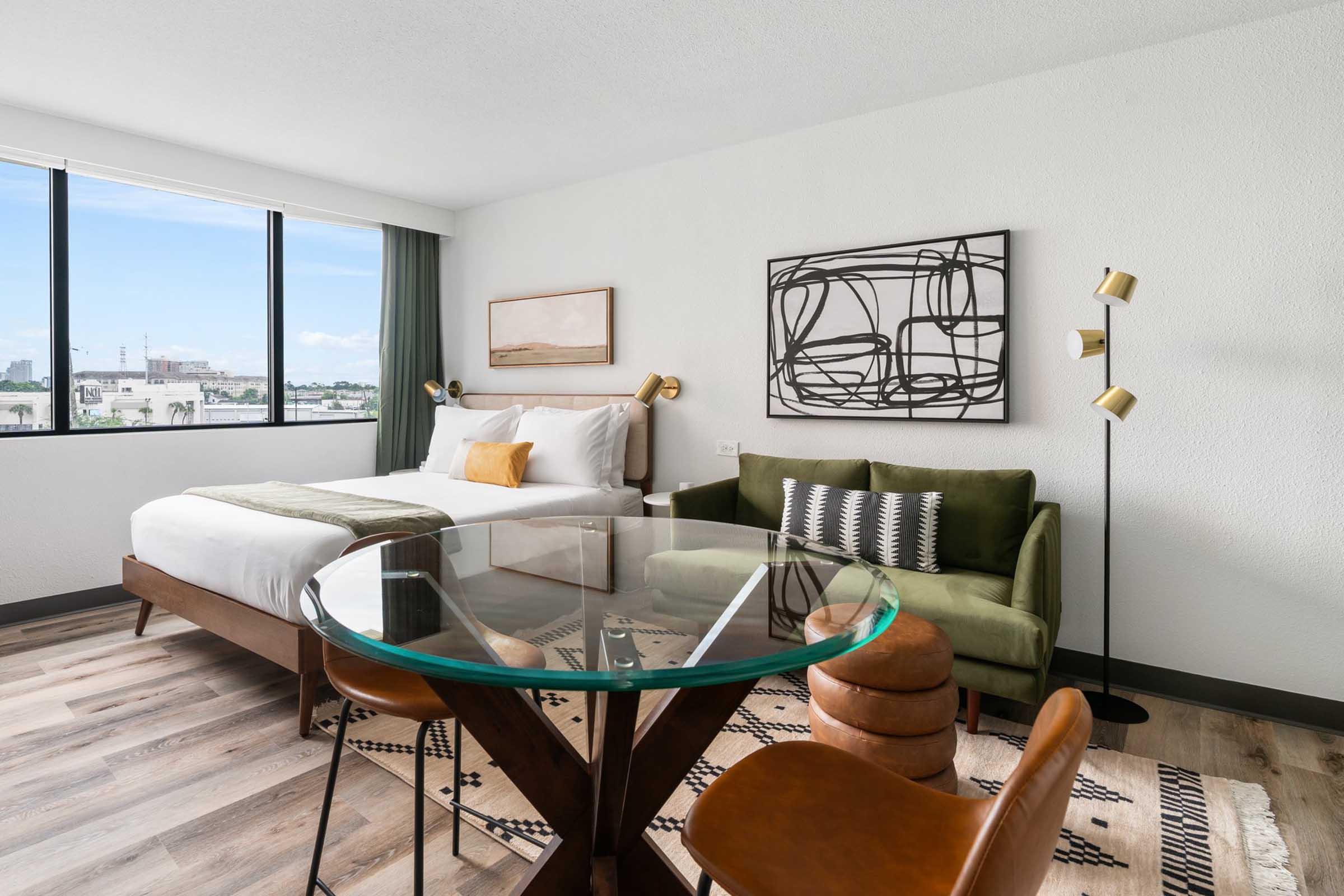 A modern hotel room featuring a king-size bed with white bedding, a green sofa, and a glass dining table. Large windows allow natural light, offering a view of the outdoors. The decor includes abstract art on the wall and a stylish rug on the floor.