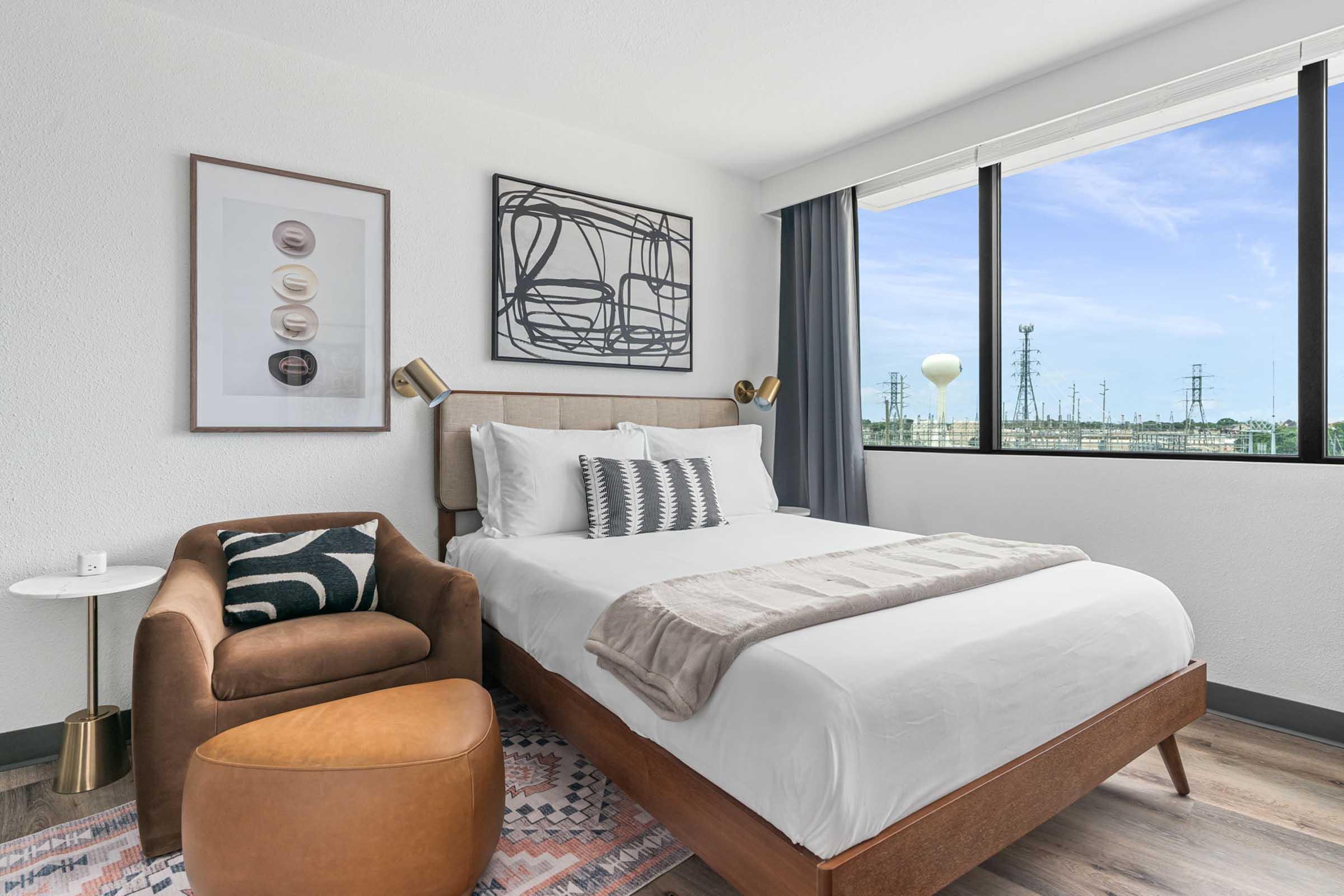 A modern hotel room featuring a queen-sized bed with white linens, a stylish armchair, and a small side table. The room has large windows with a view of power lines, framed artwork on the wall, and a cozy atmosphere enhanced by warm tones and contemporary decor.