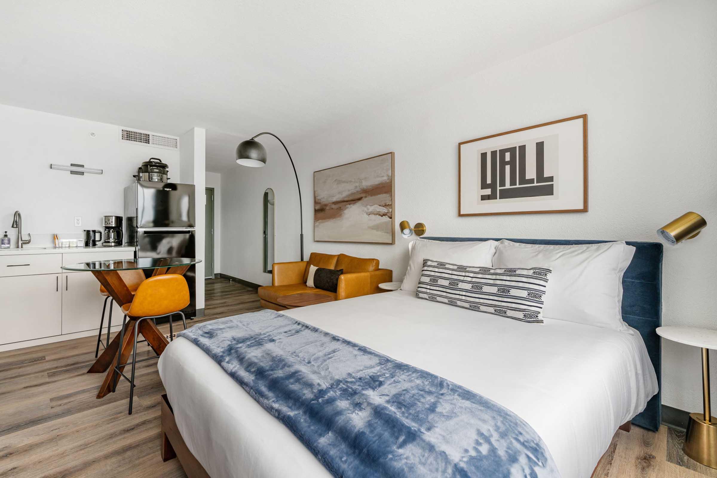 A modern hotel room featuring a queen bed with a blue throw, a small dining area with a stylish table and yellow chairs, a cozy sofa, and a kitchenette. Large artwork is displayed on the walls, and the space has a bright, inviting atmosphere with wood flooring.