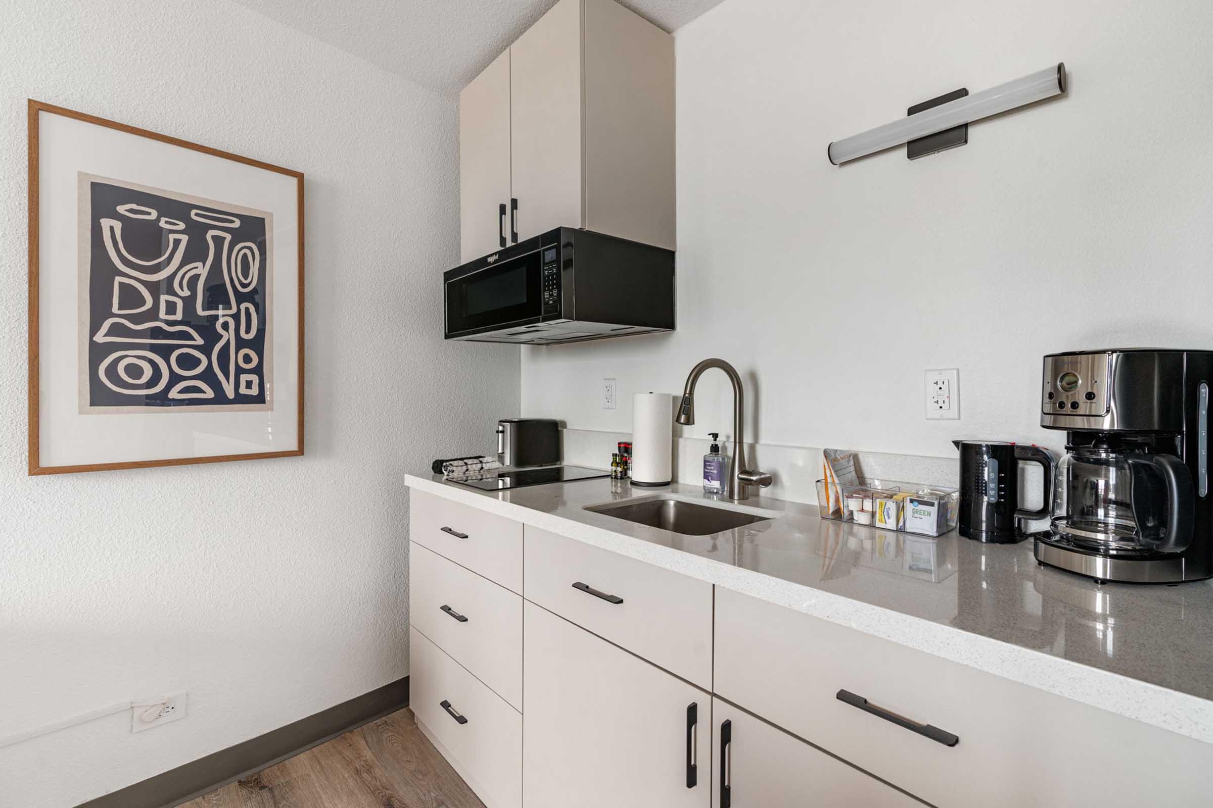 A modern kitchen area featuring light-colored cabinetry, a countertop with a sink, a microwave above the counter, a coffee maker, and a toaster. A piece of abstract wall art adds color to the space. The floor is wood-like, and the walls are painted white, creating a clean and contemporary atmosphere.