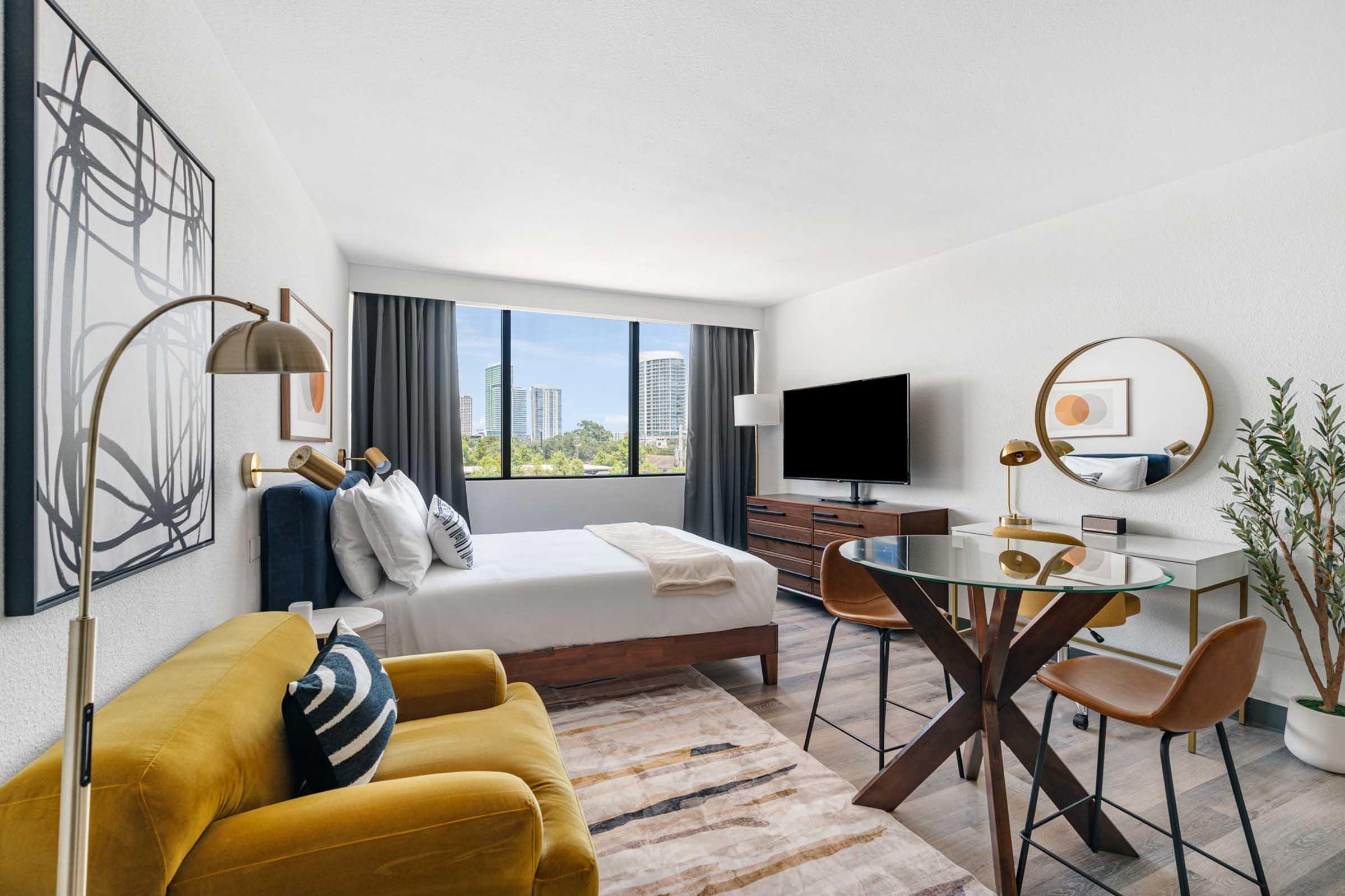 A modern hotel room featuring a cozy bed, stylish yellow sofa, circular glass dining table with chairs, and a large window with city views. Decor includes abstract artwork and greenery, enhancing the room's contemporary aesthetic. A flat-screen TV is mounted on the wall for entertainment.