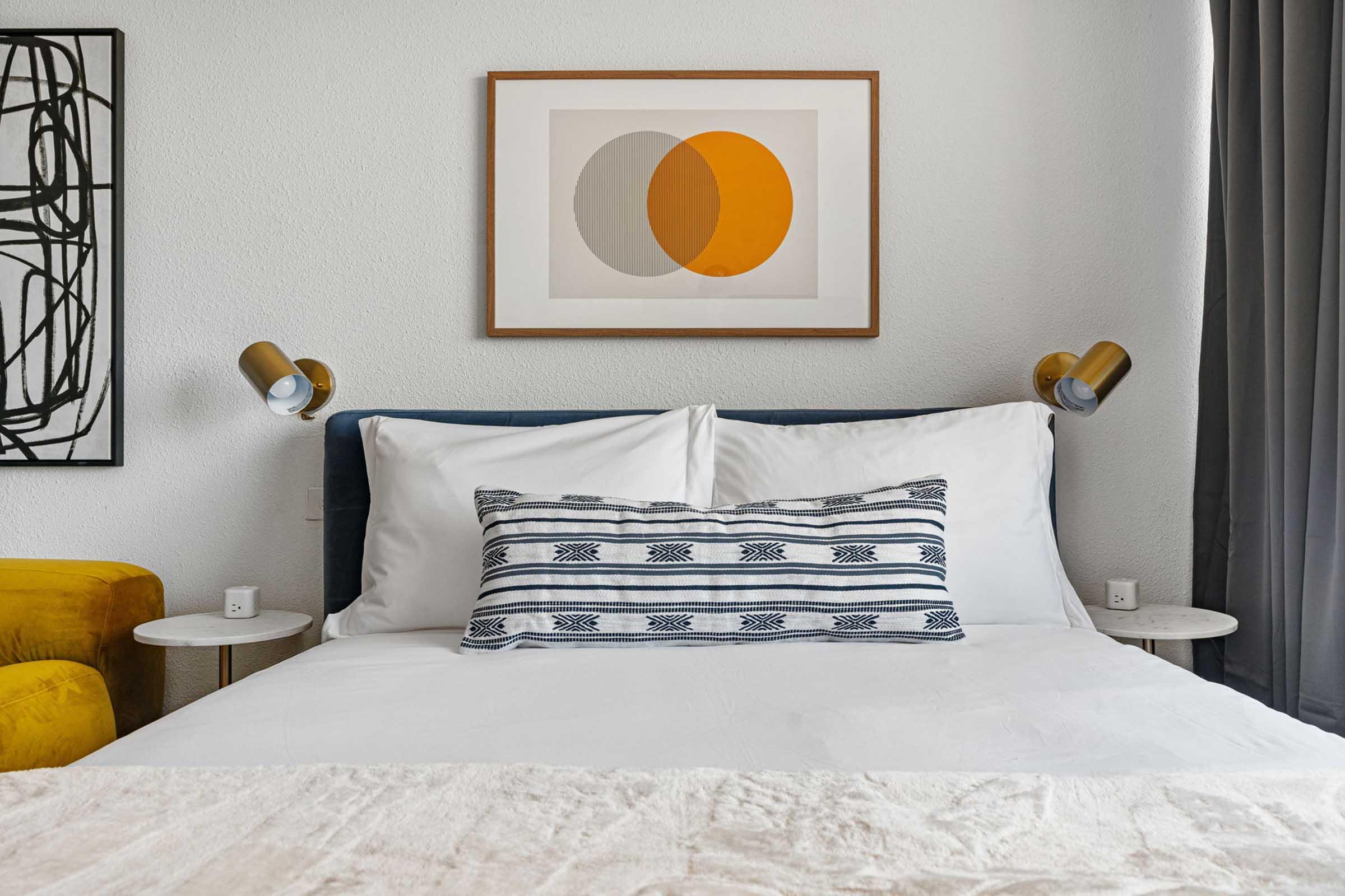 A cozy bedroom featuring a neatly made bed with white bedding and a decorative pillow. Above the bed hangs a framed abstract art piece with two overlapping circles in gray and orange. There are two stylish bedside tables with lamps, and a mustard-yellow accent chair adds a pop of color to the room.