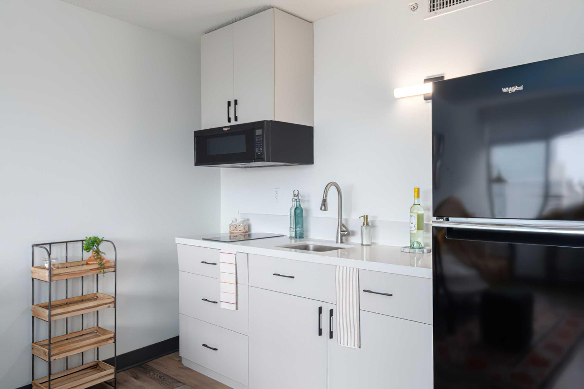 Modern kitchen featuring a sleek black refrigerator, built-in microwave, white cabinets, and a double sink with a stylish faucet. A wooden cart with shelves sits in the corner, adorned with decorative items, while light-colored countertops enhance the contemporary look of the space.