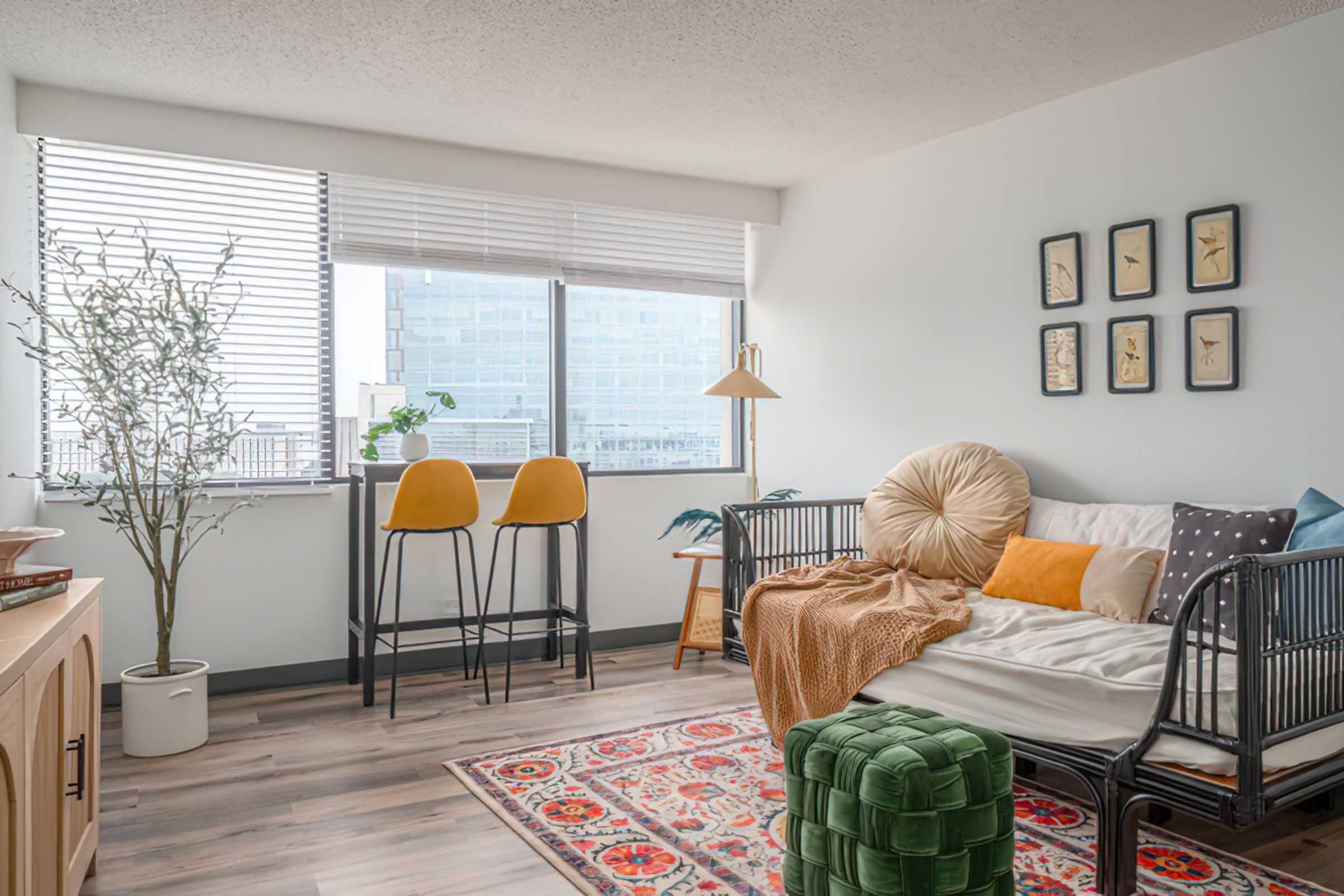 A cozy living room with a sofa adorned with colorful pillows and a throw blanket. Two yellow bar stools are positioned at a counter near a large window with blinds, allowing natural light to fill the space. Decorative art hangs on the wall, and a small green ottoman is placed on a patterned rug.