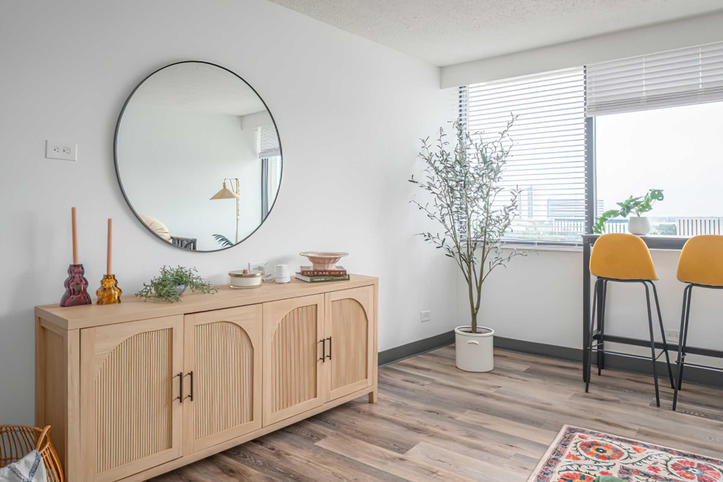 A modern room featuring a light wooden sideboard with arched doors, a large round mirror on the wall, a decorative plant in a pot, and two stylish yellow barstools nearby. Natural light streams in through window blinds, enhancing the contemporary decor and wooden flooring.