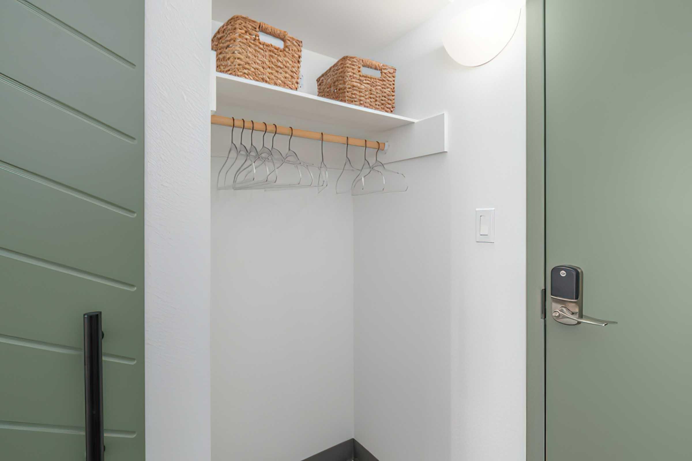 A clean and organized closet featuring a green door, a shelf with two woven baskets, and several hangers on a rod. The walls are white, giving a bright and minimalist appearance.