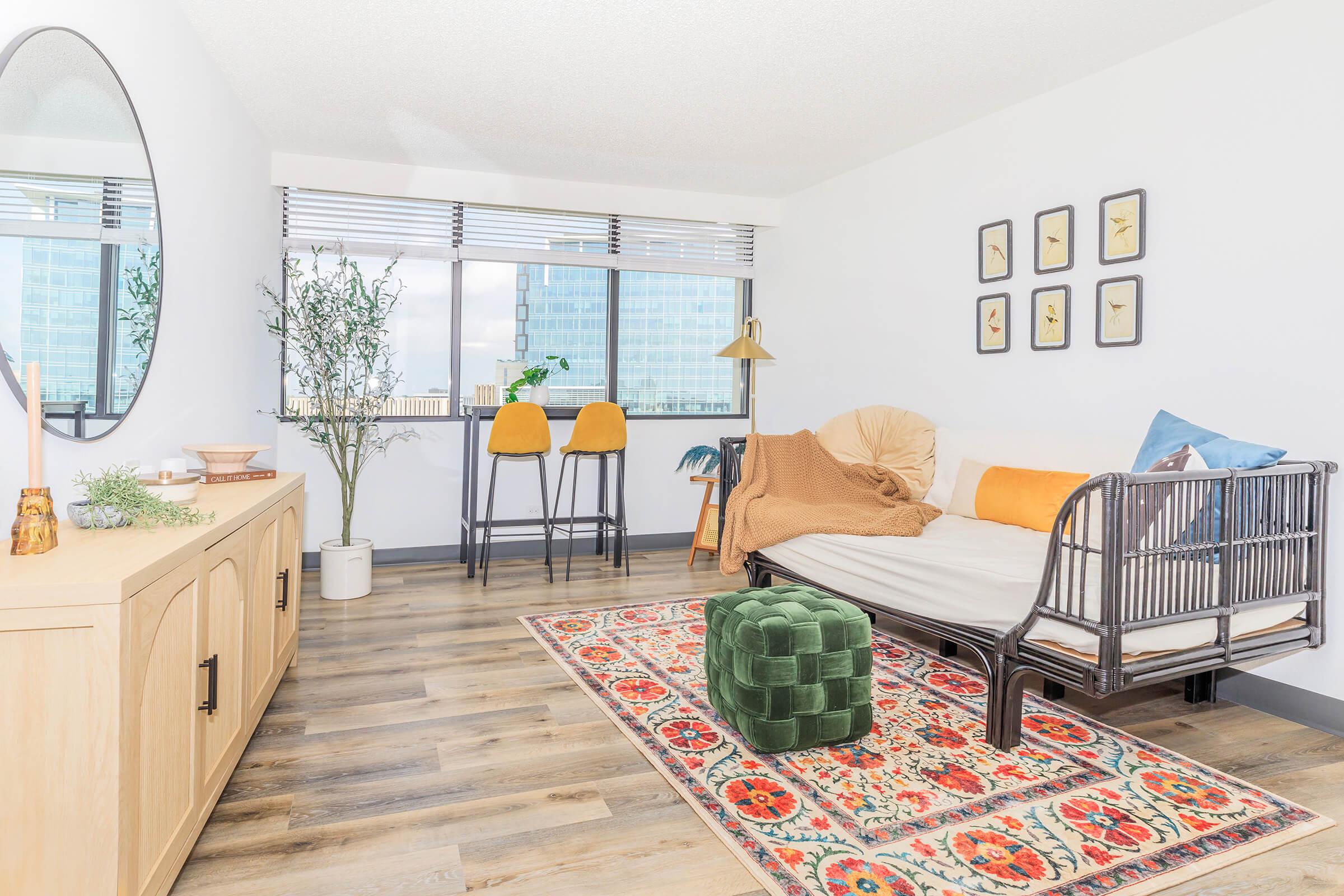 A bright and modern living space featuring a stylish couch with an orange pillow, a green ottoman, and a large round mirror above a wooden sideboard. The room has a warm color palette with a decorative rug, plants, and bar stools by a window showcasing city views. Natural light fills the area, creating an inviting atmosphere.