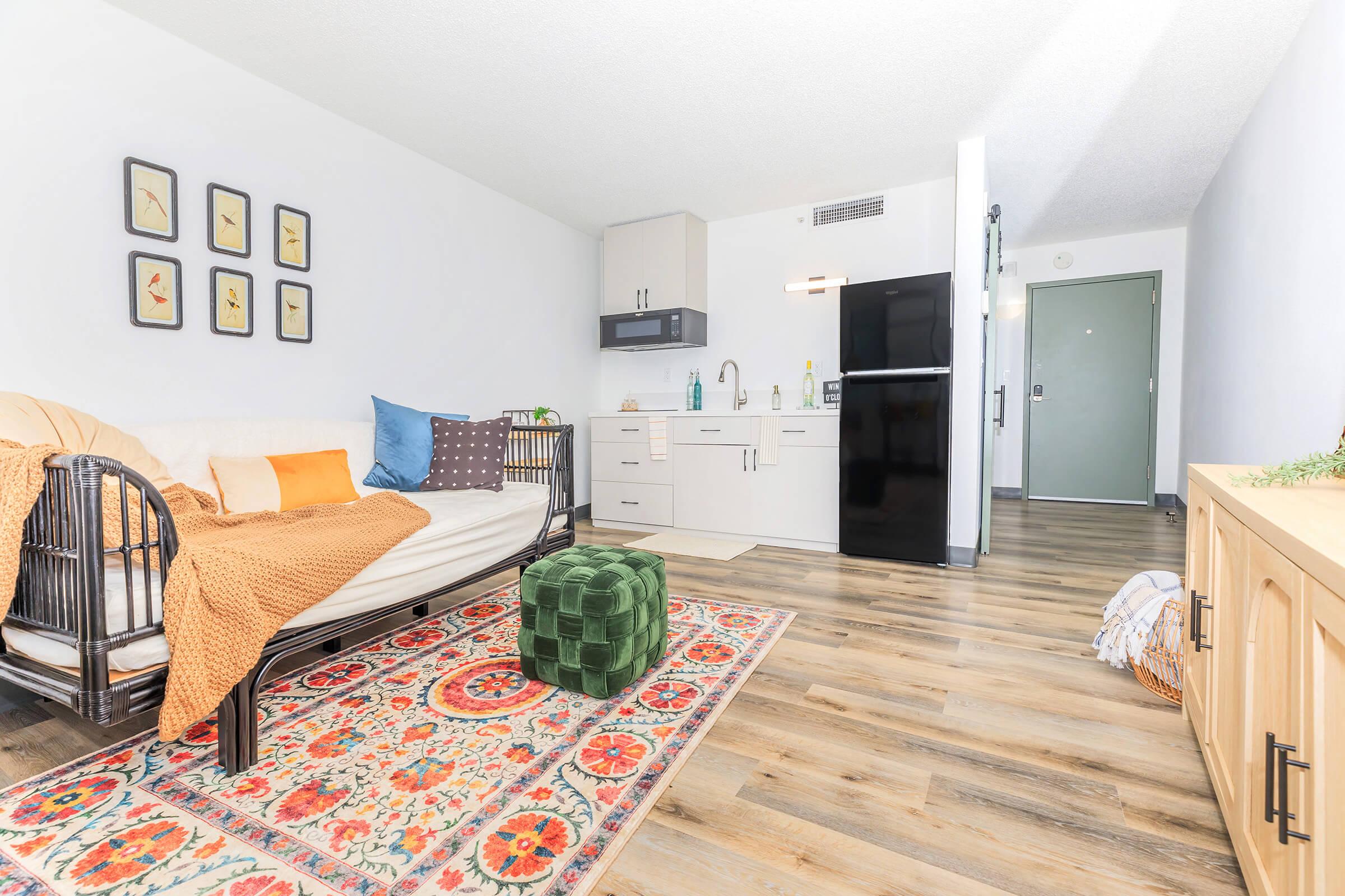A cozy living space featuring a daybed adorned with a brown throw and colorful pillows, situated on a decorative rug. There is a small kitchen area with modern appliances, including a black refrigerator. Natural light streams in from a window, enhancing the bright, inviting atmosphere of the room.