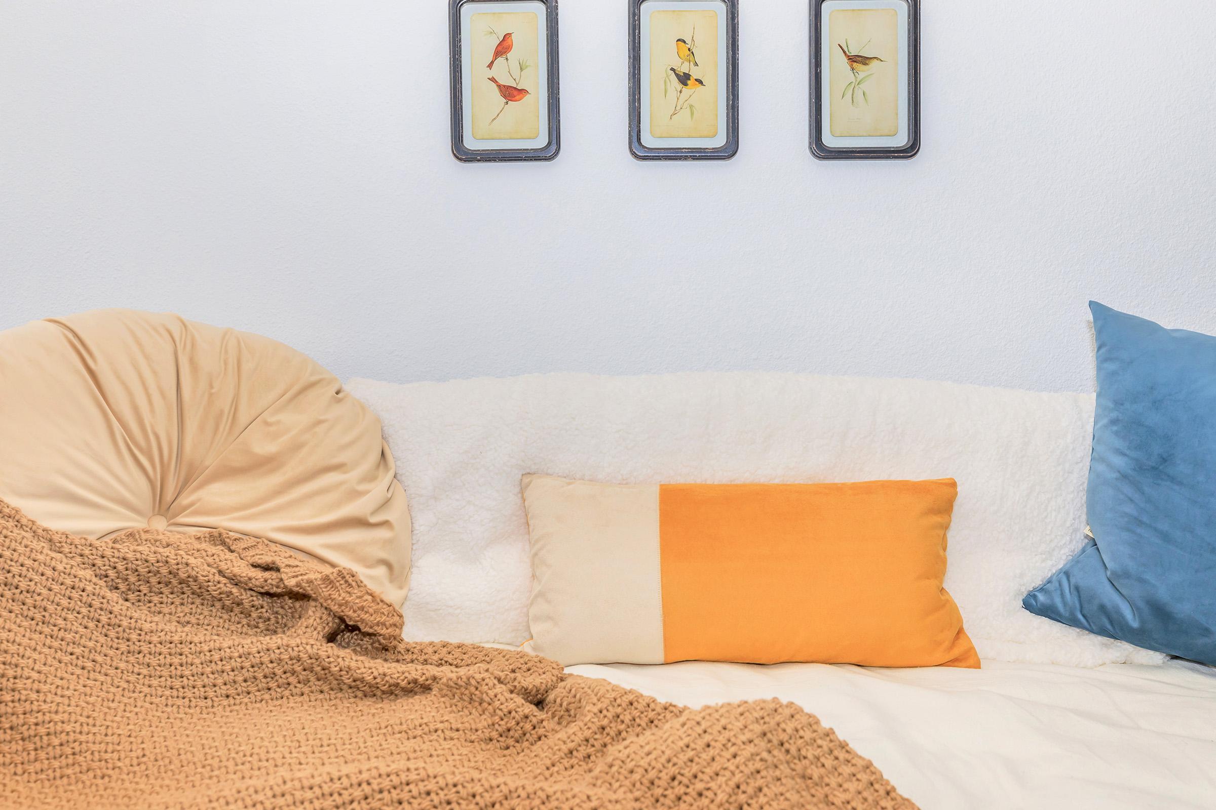 A cozy couch adorned with a fluffy white throw and a textured brown blanket. The couch features a rectangular orange and cream pillow and two decorative pillows in soft blue and beige. Above the couch, three framed bird illustrations hang on a light-colored wall, adding a touch of warmth to the space.