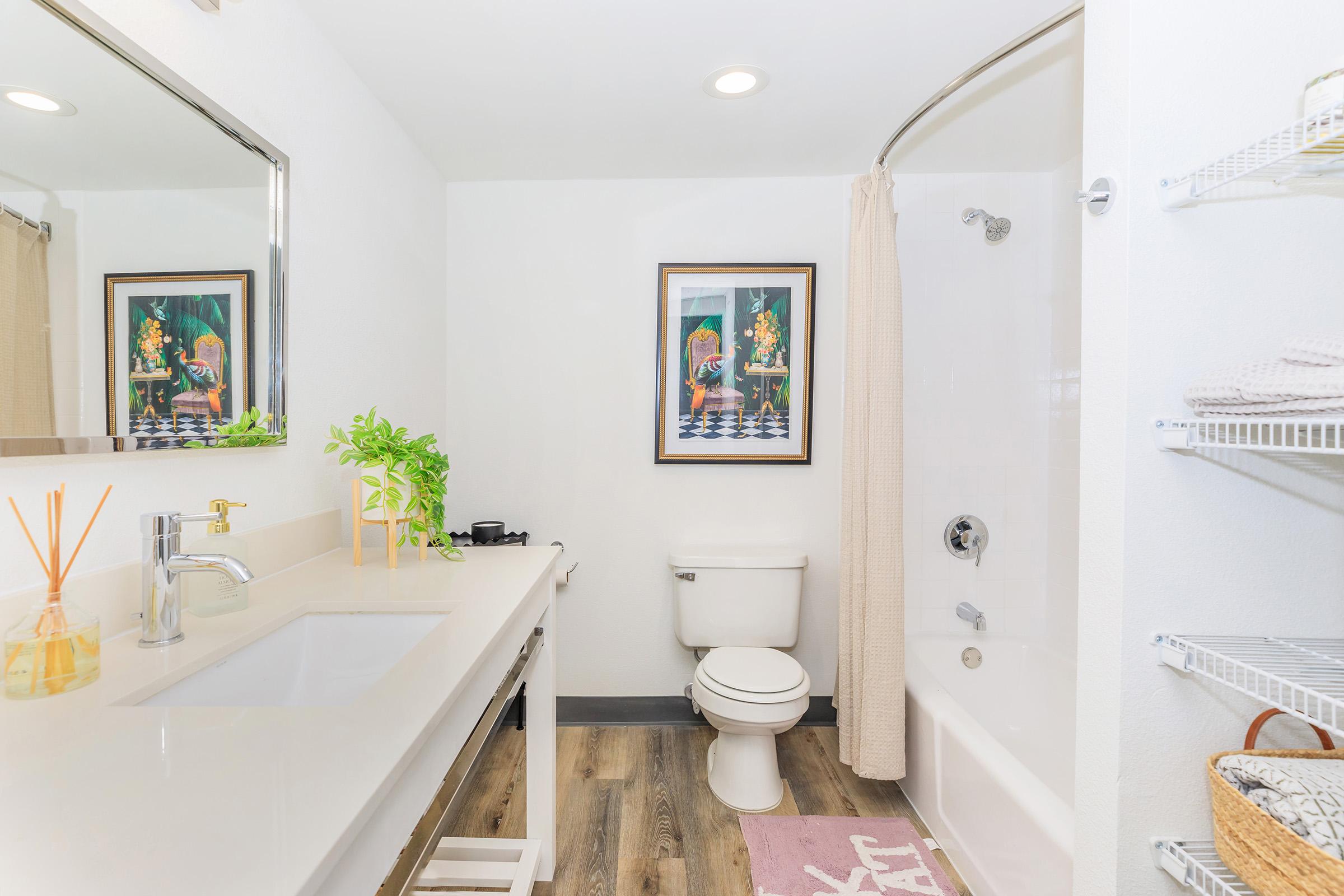 A modern bathroom featuring a white sink with a large mirror above it, a toilet, and a bathtub with a shower curtain. The walls are painted white, and there is a decorative plant on the counter. A framed piece of artwork hangs on the wall, and there is a storage rack with neatly organized items.