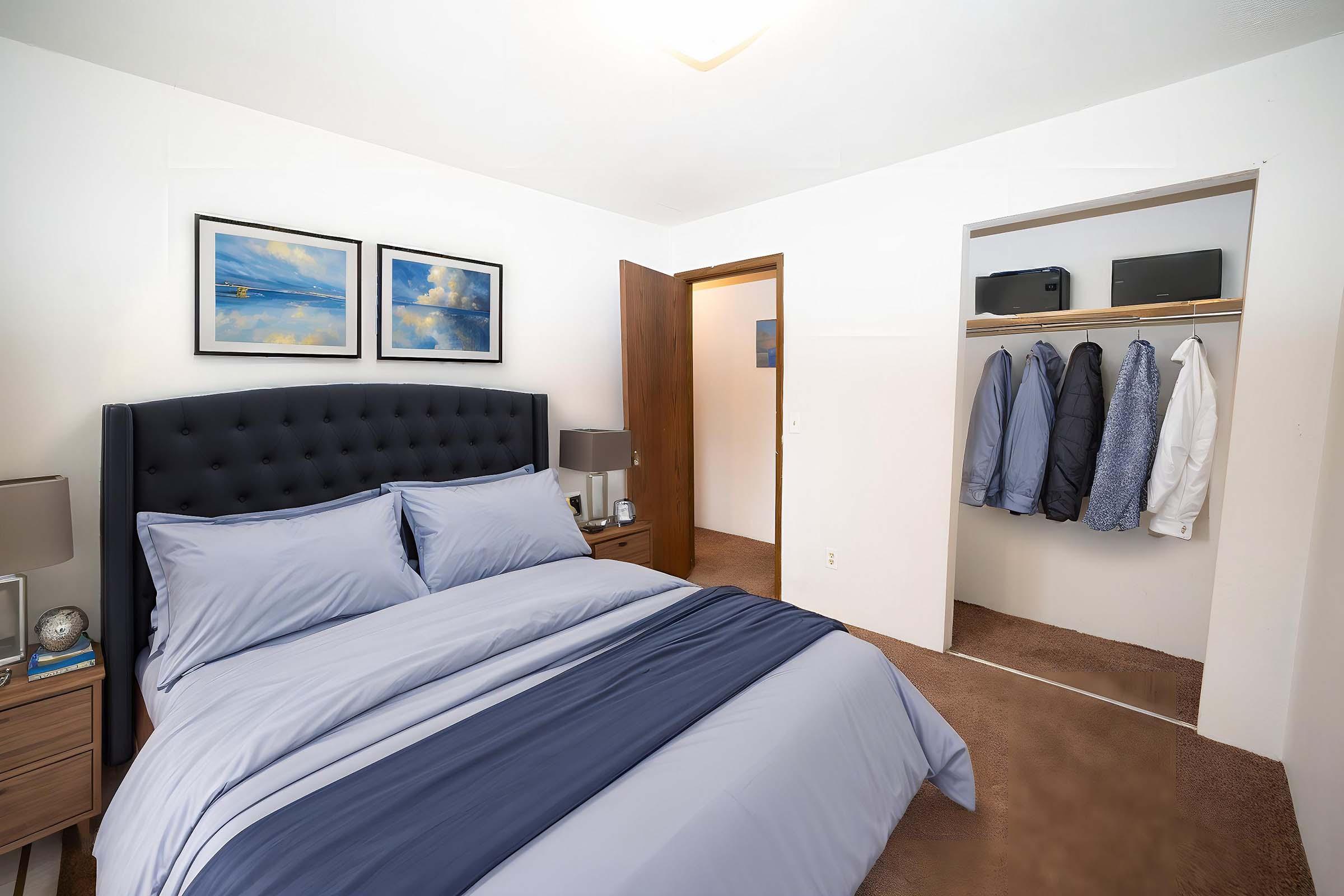 A neatly arranged bedroom featuring a dark blue upholstered bed with matching linens. Two framed landscape pictures hang above the bed. To the right, a closet with several neatly hung coats is visible, and a bedside table with a clock and lamp sits on the left. The room has neutral wall colors and carpeted flooring.