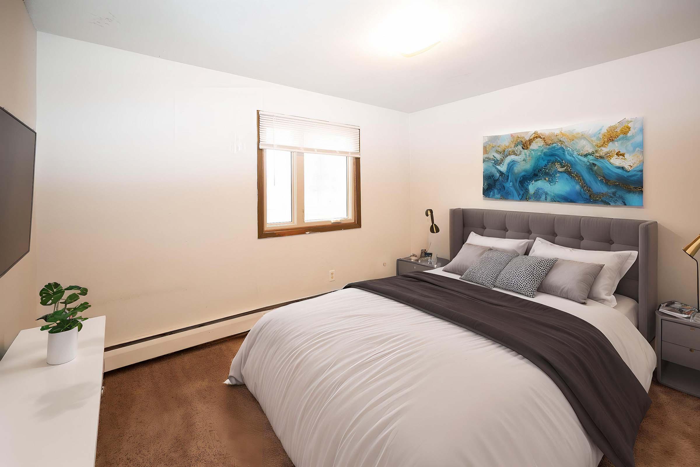 A cozy bedroom featuring a neatly made bed with gray and white bedding, a decorative painting above the headboard, a window with blinds, and a modern bedside lamp. A small plant sits on a white dresser opposite the bed, and a television mounted on the wall completes the inviting space.