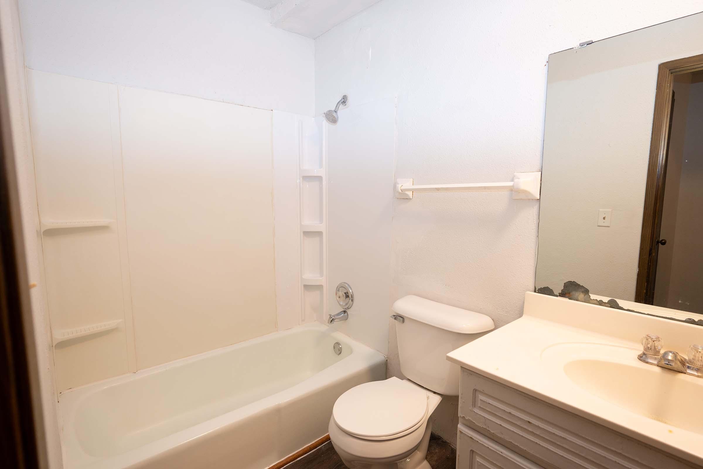 A small, simple bathroom featuring a white bathtub, toilet, and a sink with a countertop. The walls are painted white, and there is a mirror above the sink. A showerhead is installed above the tub, and there are built-in shelves on the wall next to the bath.