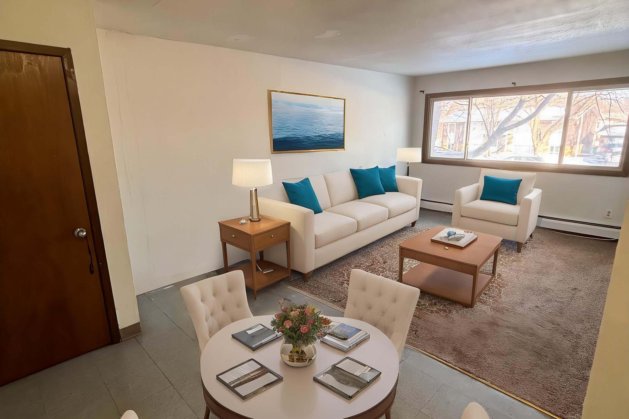 A cozy living room featuring a white sofa with turquoise cushions, a wooden coffee table, and a dining area with a round table set for four. Large windows allow natural light, and there’s a piece of wall art depicting water. The space has a warm and inviting ambiance.