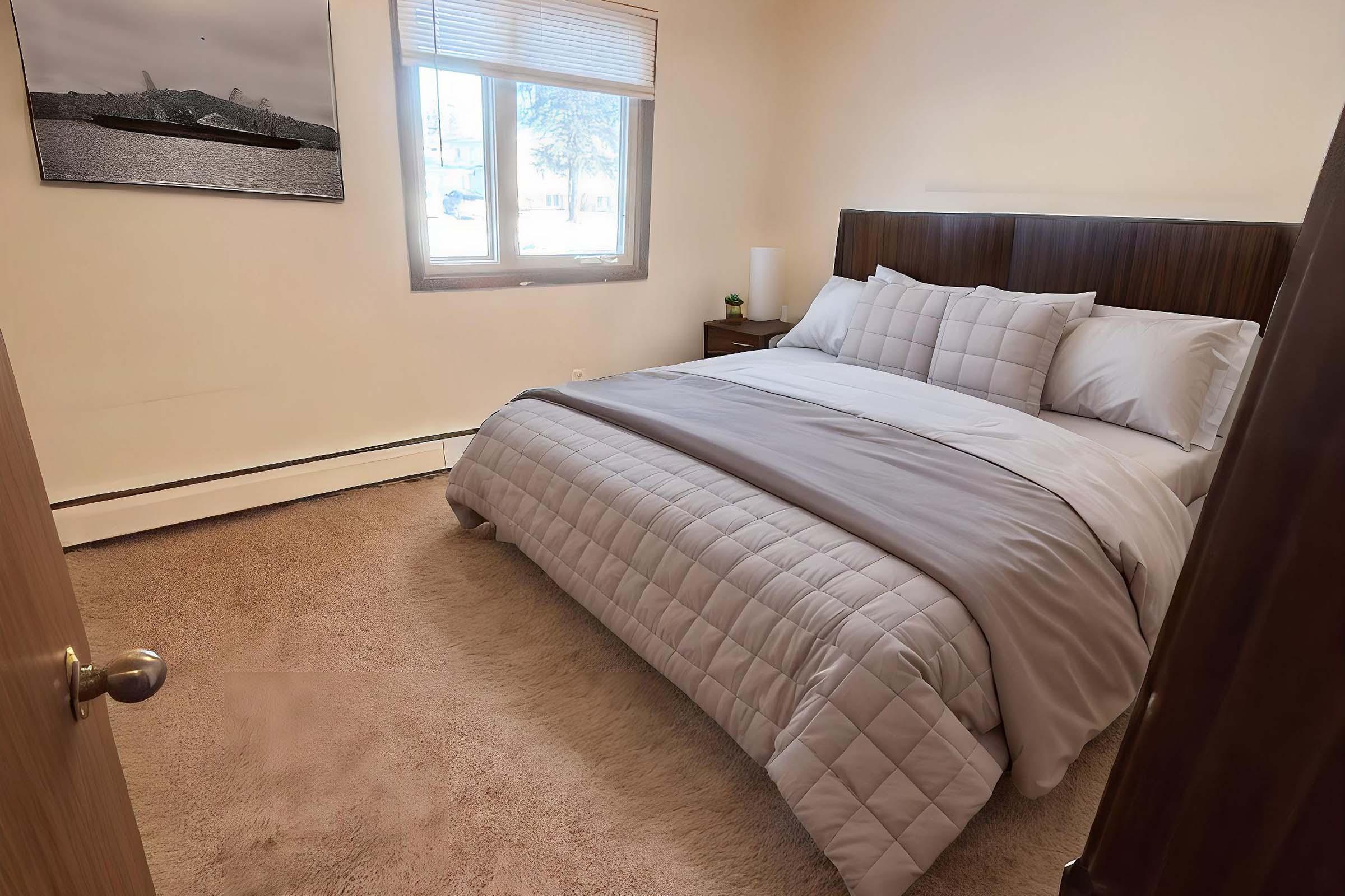 A cozy bedroom featuring a large, neatly made bed with a light gray quilt and multiple pillows. There is a bedside table with a lamp beside the bed, and a window allowing natural light into the room. A framed black-and-white photograph is hung on the wall, and the floor is covered with soft carpeting.