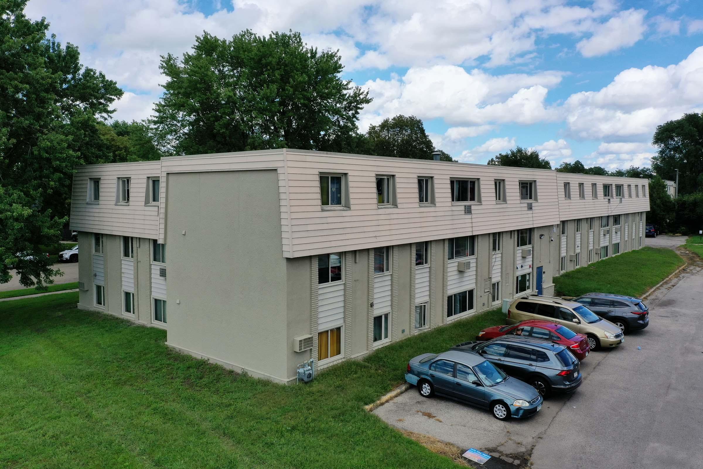 A two-story apartment building with a light-colored exterior and several windows. The grass is green, and a few cars are parked along the side of the building. The sky is partly cloudy with blue patches visible.