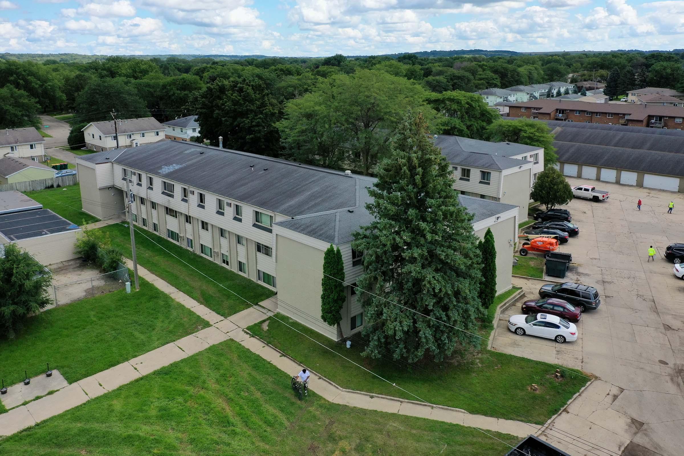 Aerial view of a multi-story apartment complex surrounded by greenery. The building features multiple windows and a flat roof, with trees nearby and parking spaces filled with cars. A pathway runs along the building, leading to the entrance area. In the background, additional residential structures can be seen under a partly cloudy sky.