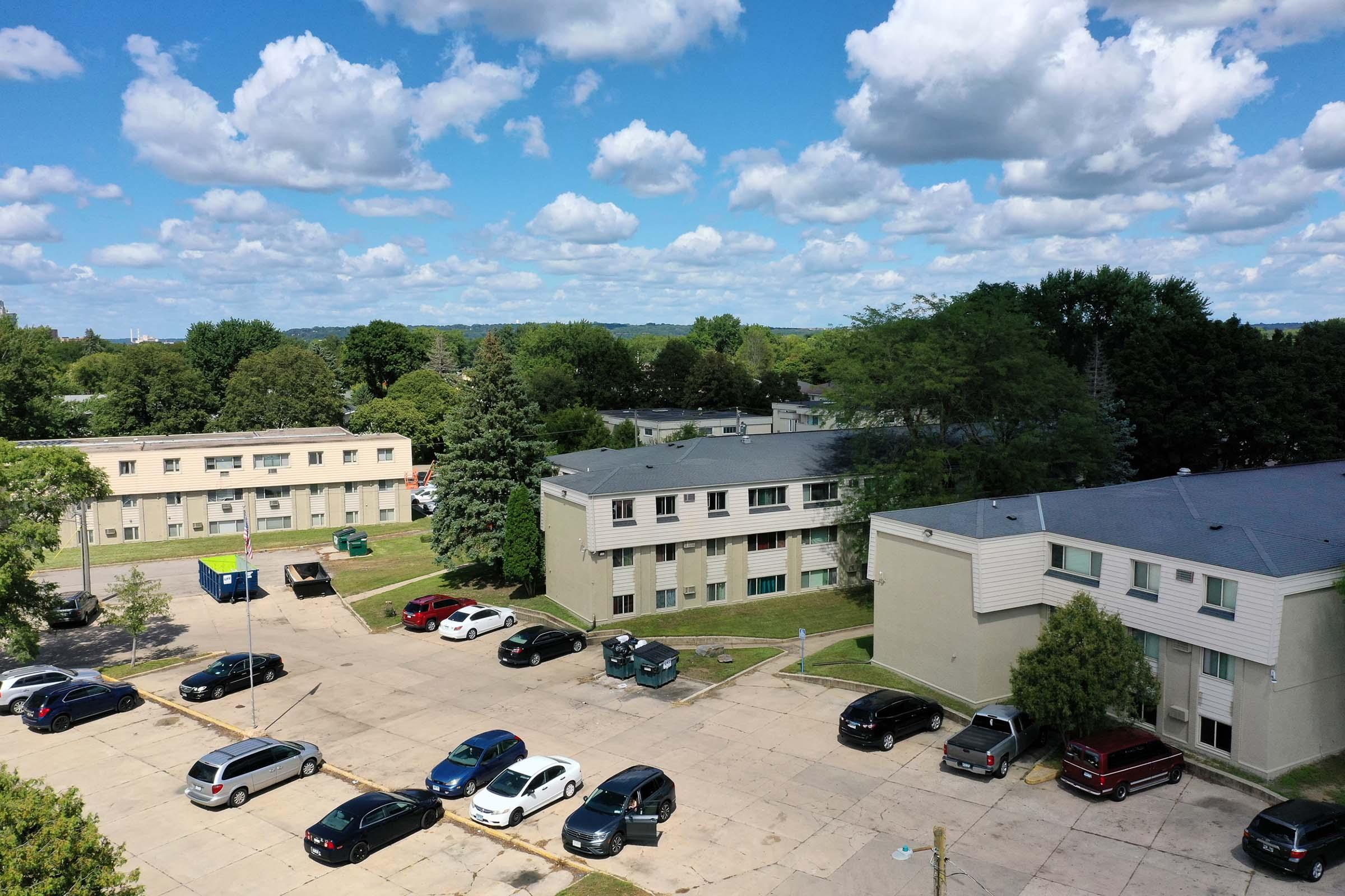 Aerial view of a residential area featuring multiple apartment buildings and a parking lot filled with cars. The scene includes green trees and blue skies with fluffy white clouds.