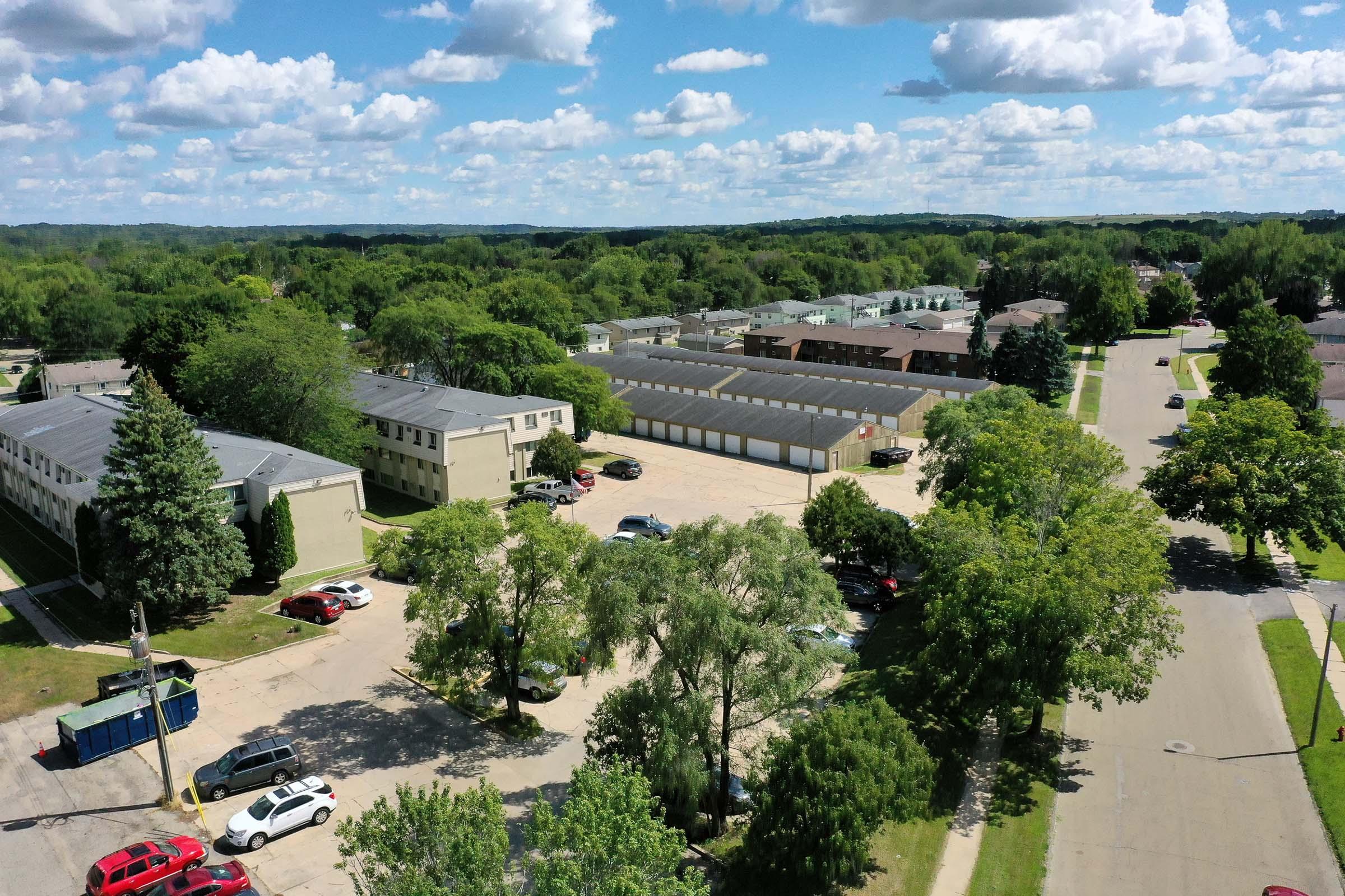 An aerial view of a suburban area featuring residential buildings, several parked cars, and a storage facility. The landscape includes trees and lush greenery, under a partly cloudy sky with expansive blue. The street is visible, lined with more homes and vegetation.
