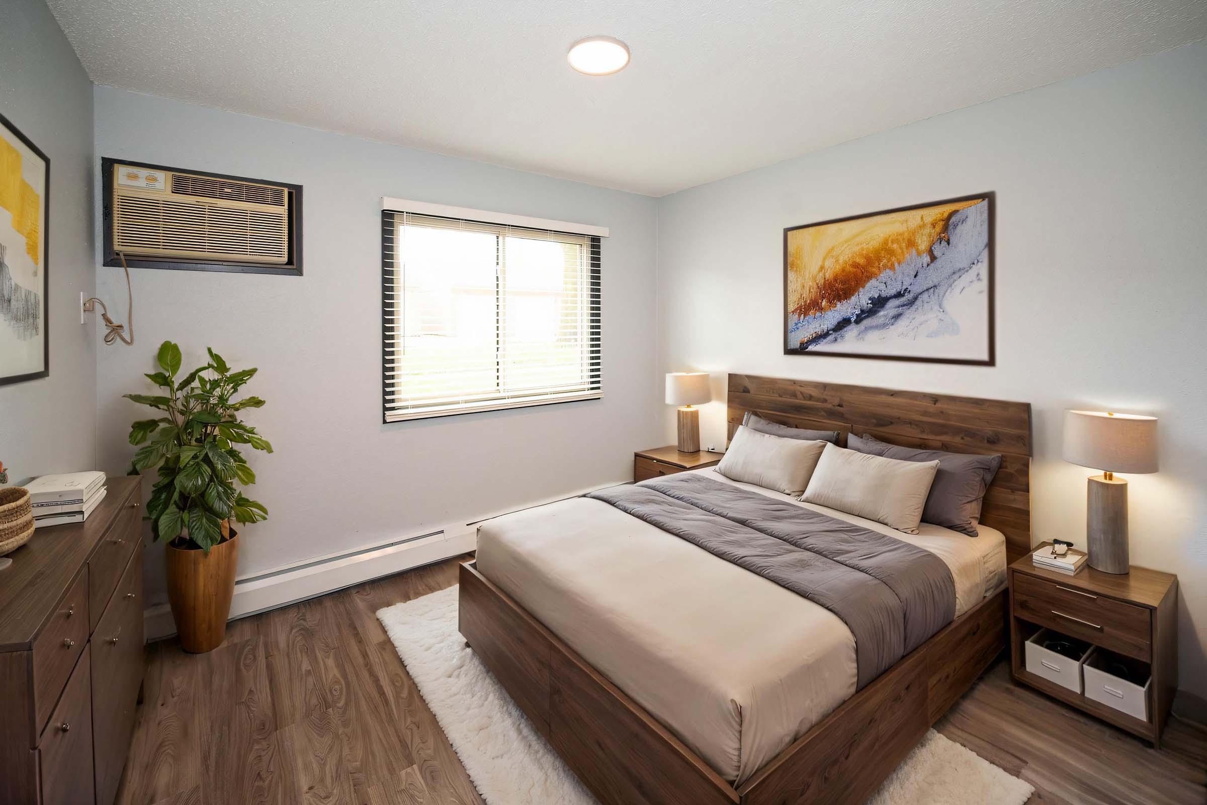 A comfortable bedroom featuring a wooden bed with gray bedding, matching nightstands with lamps, a dresser, and a potted plant. Large window allowing natural light, an air conditioning unit installed, and abstract wall art above the bed create a cozy atmosphere.