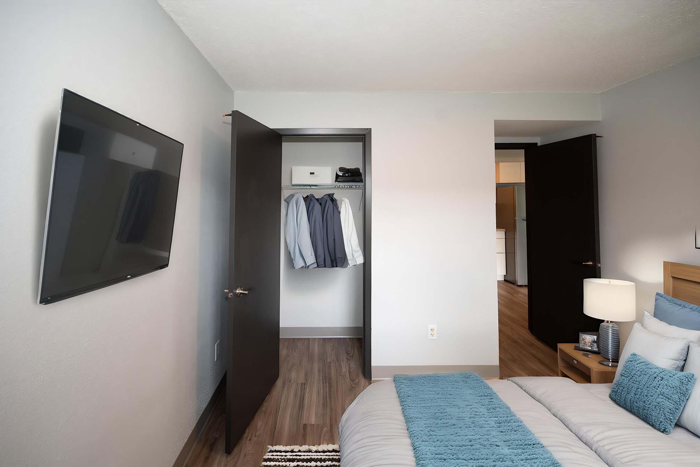 A modern bedroom featuring a neatly made bed with decorative pillows, a bedside lamp, and a wall-mounted TV. A closet with a few hanging shirts is visible in the background, along with two doors leading to other areas of the space. The walls are light gray, and the floor is wooden.