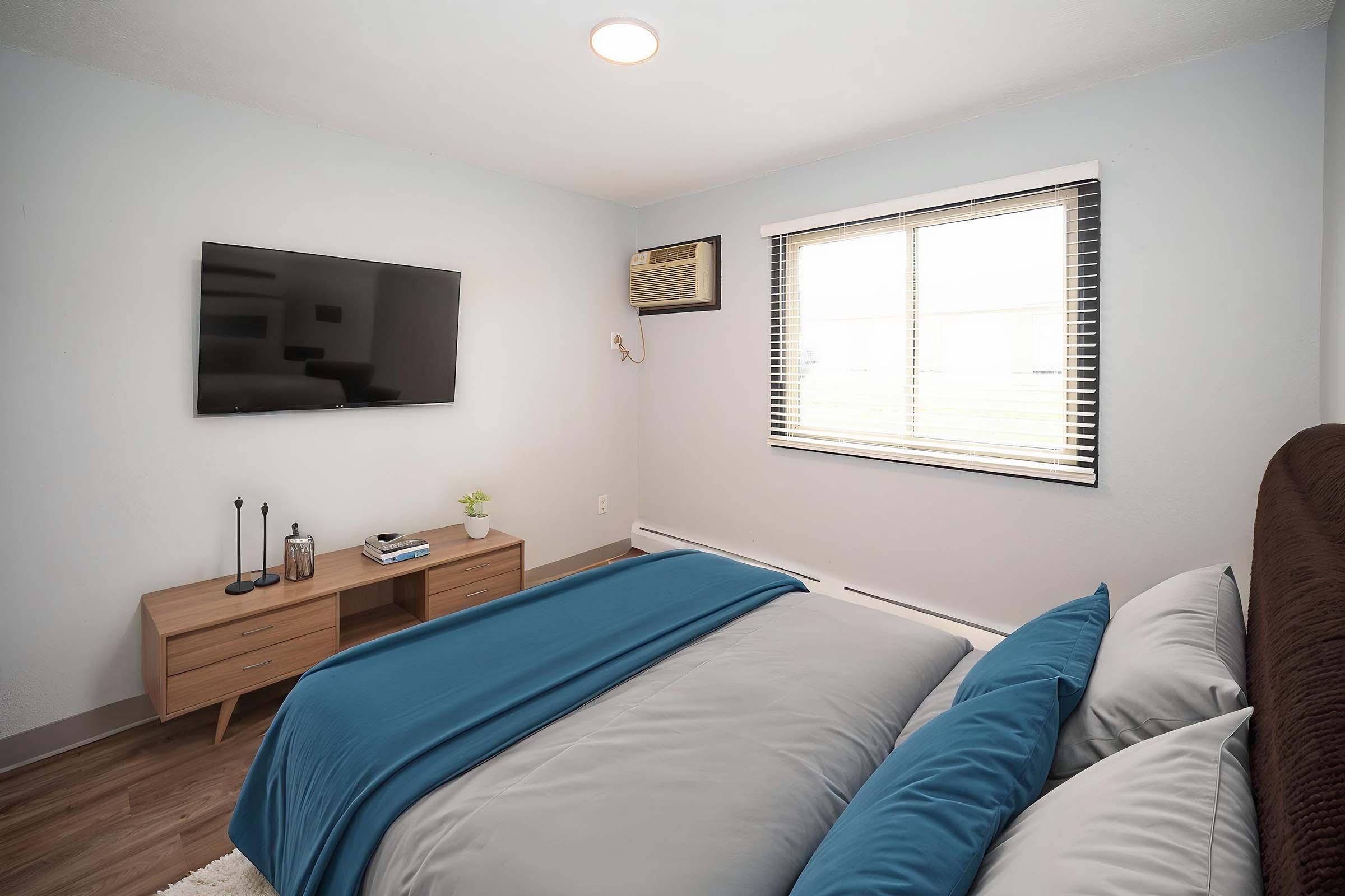 A cozy bedroom featuring a large bed with blue and gray bedding, a wooden bedside table, and a wall-mounted television. Natural light streams through a window with blinds, and there's an air conditioning unit above the window, enhancing the room's comfort.