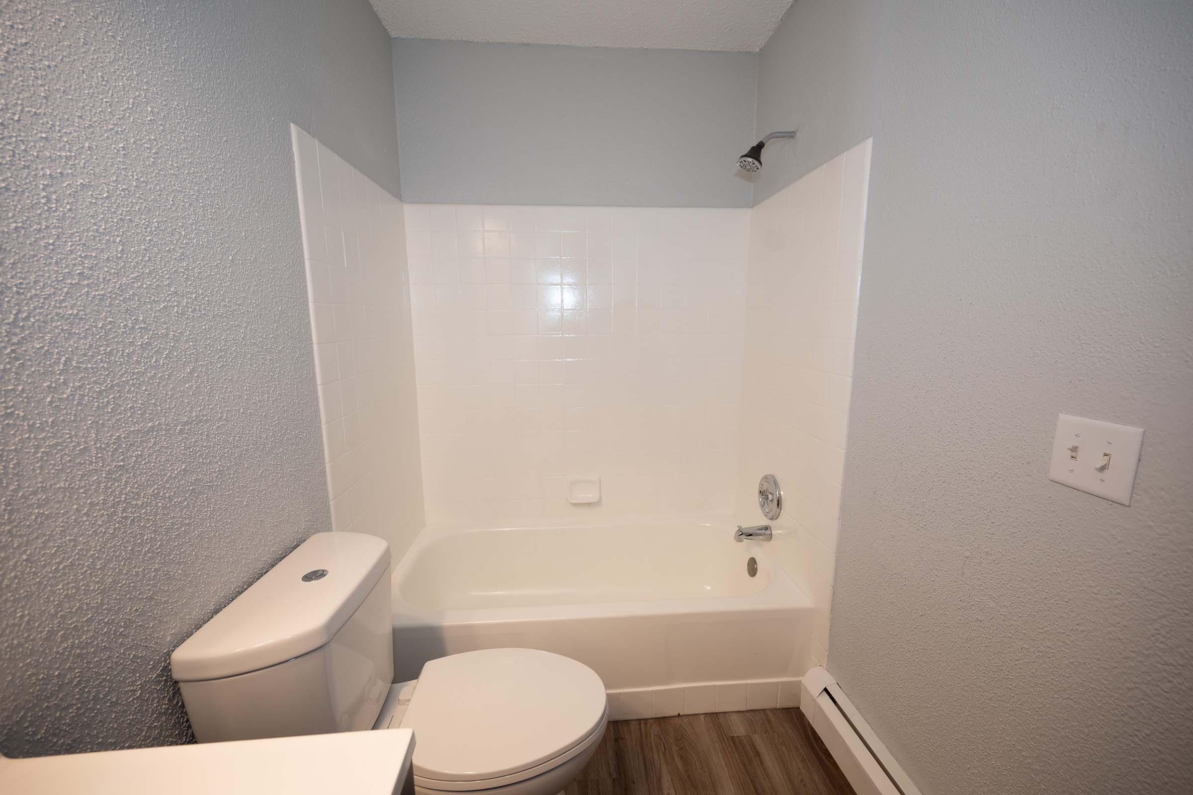 A clean and simple bathroom featuring a white bathtub with a showerhead, a modern toilet, and light gray walls. The floor has a wood-like appearance, adding warmth to the space. The overall design is minimalistic and functional, making it suitable for various styles of home interiors.
