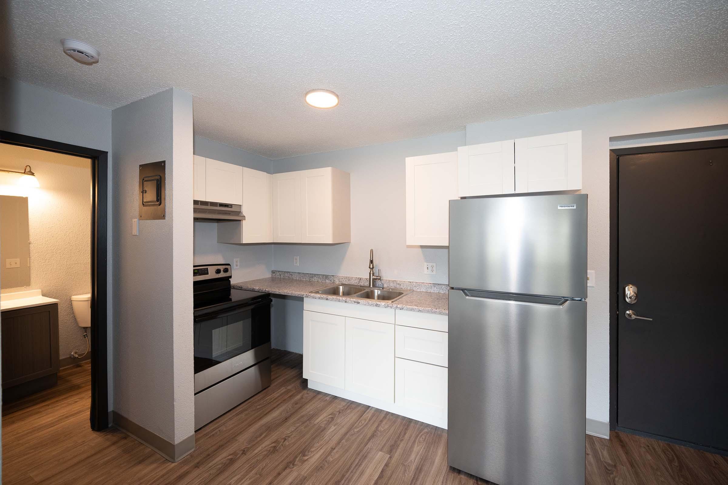A modern kitchen featuring stainless steel appliances, including a refrigerator and stove. The cabinetry is white, and there's a countertop with a sink. The space has wood-like flooring and a simple, neutral color scheme. A doorway leads to a bathroom, and a front door is visible.