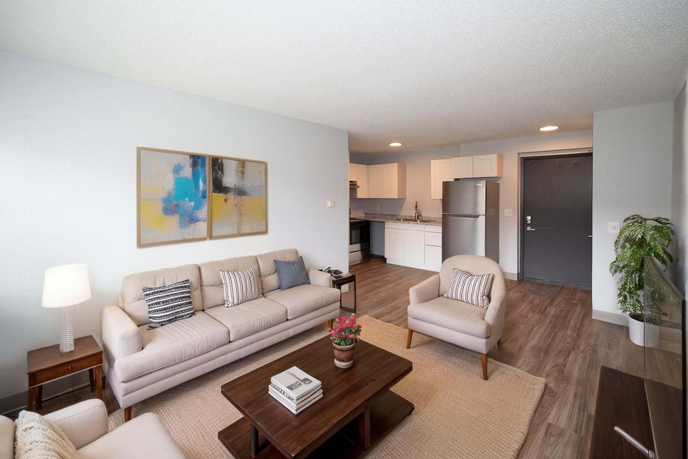 A modern living room featuring a light-colored sofa with decorative pillows, two armchairs, a wooden coffee table, and a TV on a stand. To the side, there's a dining space and a kitchen with white cabinets and appliances. A potted plant adds a touch of greenery, and artwork hangs on the wall.