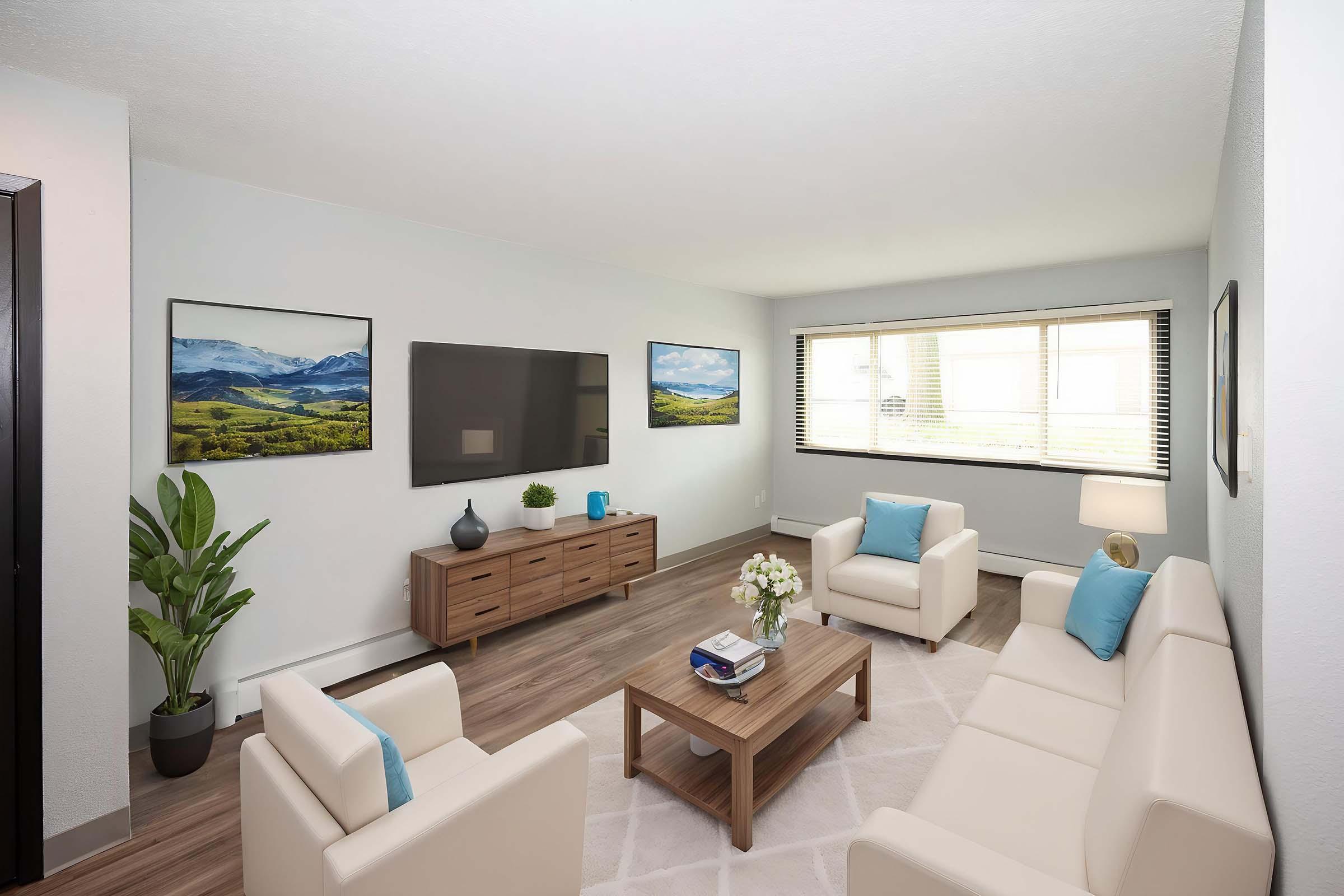 A modern living room featuring a light color scheme. It includes two white sofas, a wooden coffee table, and a TV mounted on the wall. Decorative elements include potted plants, artwork depicting landscapes, and a vase of flowers on the table, complemented by soft lighting from a lamp.