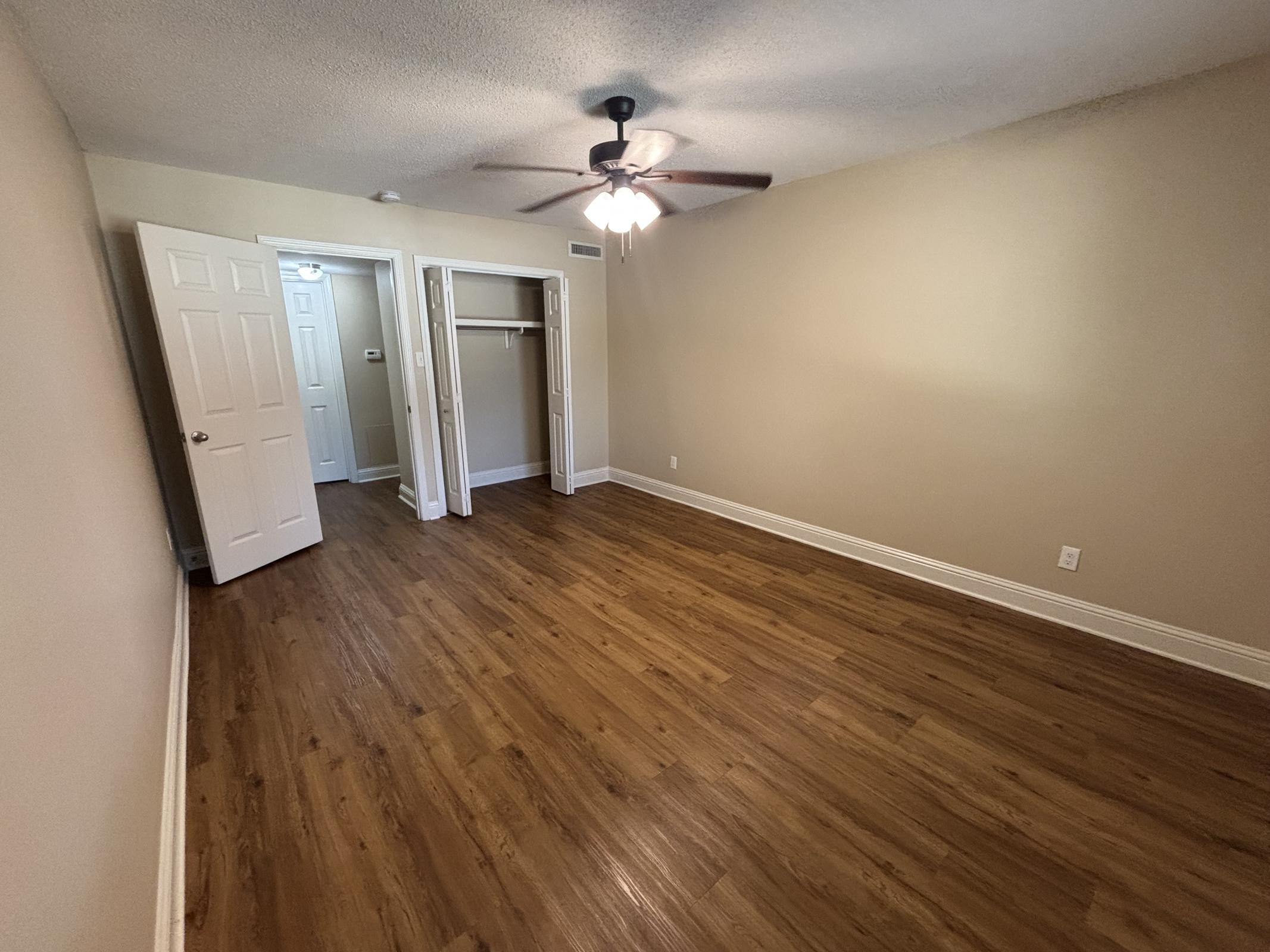 Empty room with wooden flooring, a ceiling fan, and light beige walls. Two doors on the left lead to a closet; one door is slightly open, revealing hangers. Natural light enters through a window, highlighting the spaciousness of the area.