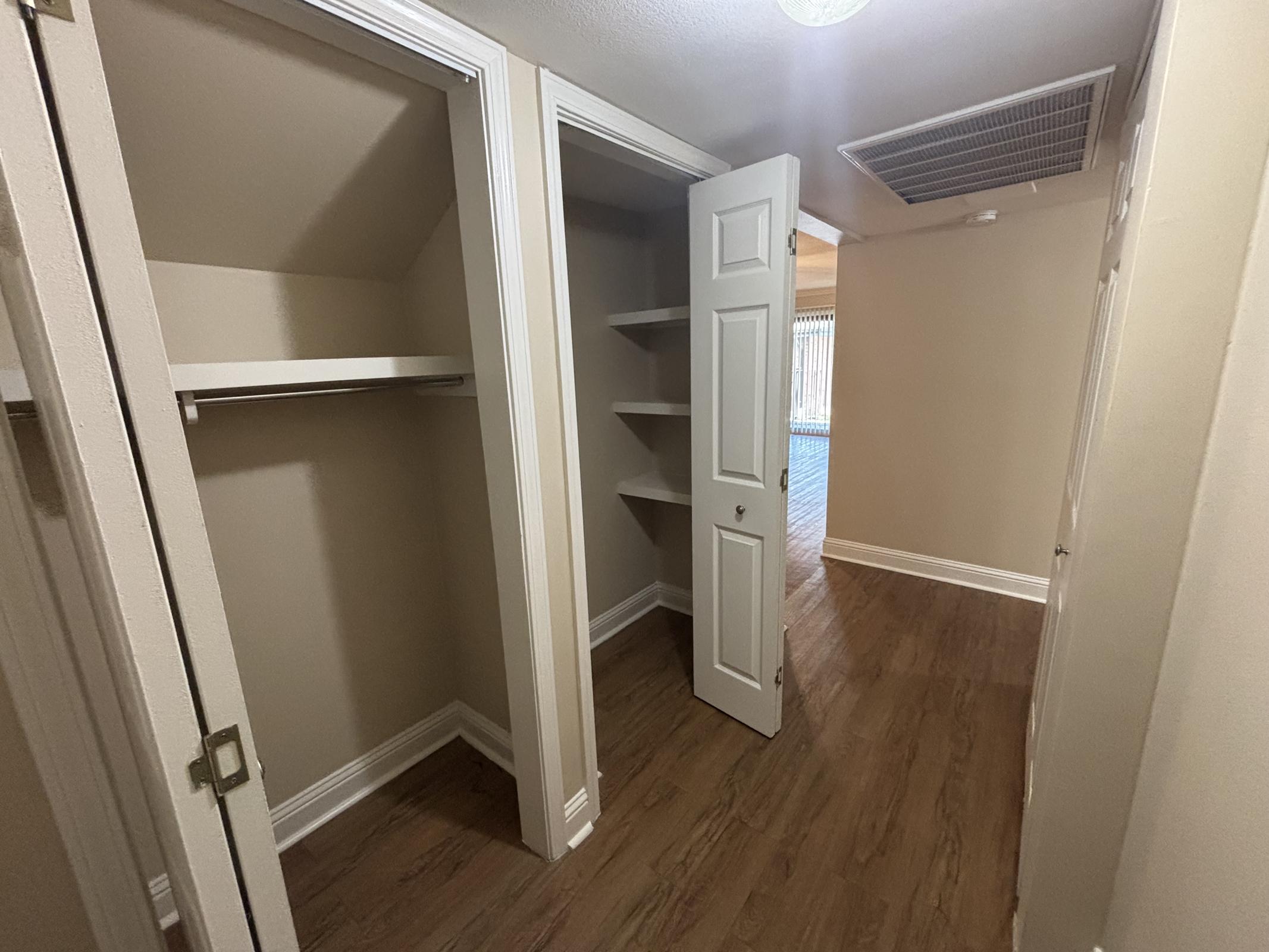 Interior view of a hallway featuring two closet doors on either side. One closet has shelving, while the other has a hanging rod. The walls are painted a light beige color, and the flooring is a wood-like laminate. Soft lighting from an overhead fixture illuminates the space.