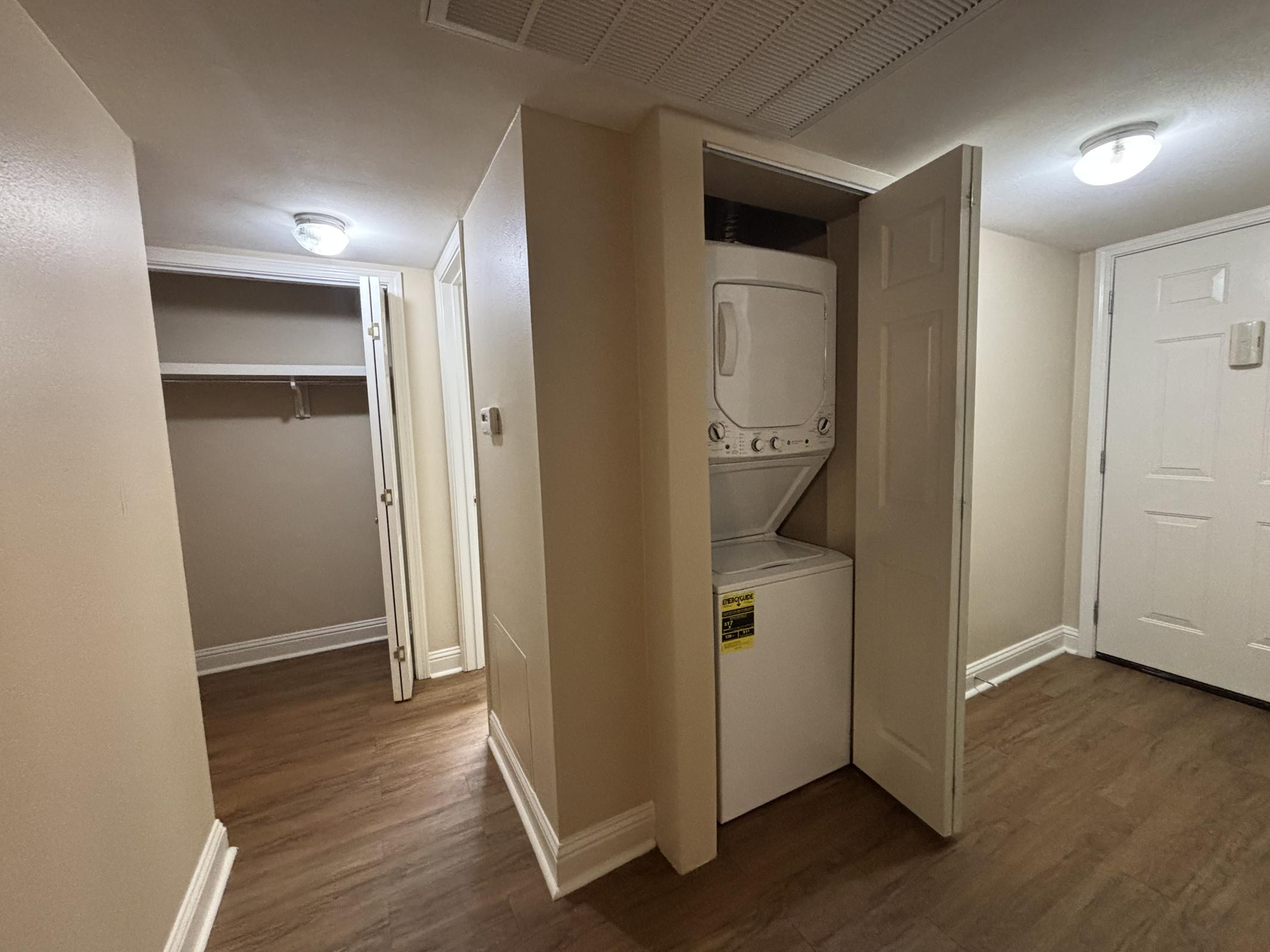 A laundry area featuring a stacked washer and dryer unit in a small, well-lit hallway. An open closet with a hanging rod is visible to the left, and a closed door is seen on the right. The walls are painted beige, and the flooring is a wood-like finish. Ceiling lights provide illumination.
