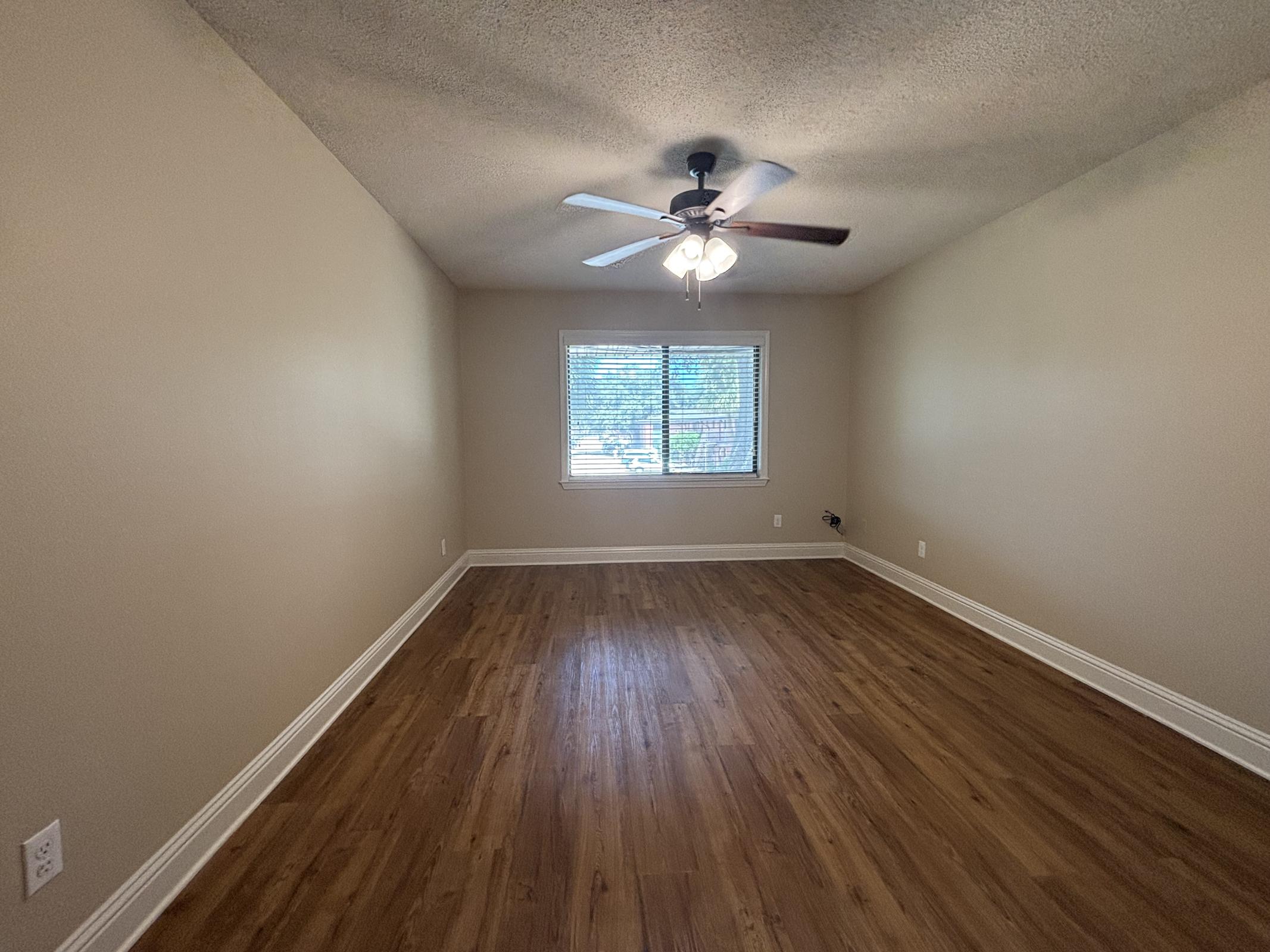 A spacious room with light beige walls and a ceiling fan. There are two windows allowing natural light to enter, and the floor is made of hardwood. The room is uncluttered and empty, providing a blank space for arrangement.
