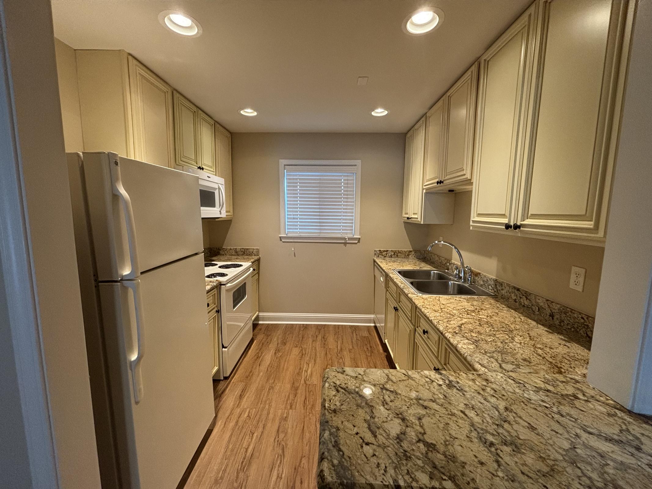 A modern kitchen featuring light-colored cabinets, granite countertops, a white refrigerator, a stove with an oven, a microwave, and a sink. The space has warm wood flooring and an adjacent window allowing natural light. Ceiling lights illuminate the room.