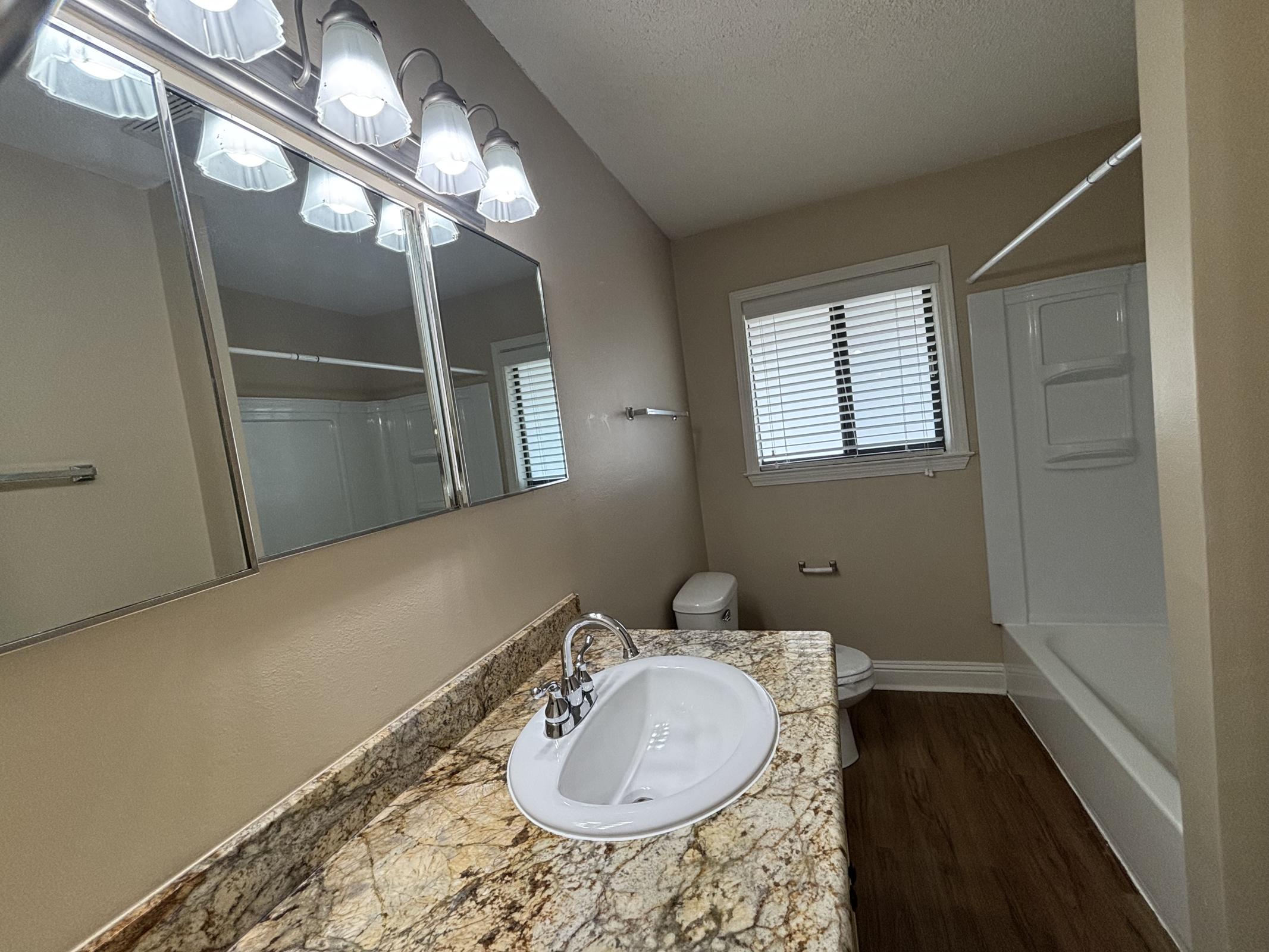 A well-lit bathroom featuring a large mirror with multiple light fixtures, a sink with a stylish faucet, and a granite countertop. It includes a toilet and a bathtub with a shower, all set against light-colored walls and wooden flooring. Natural light enters through a window with blinds.