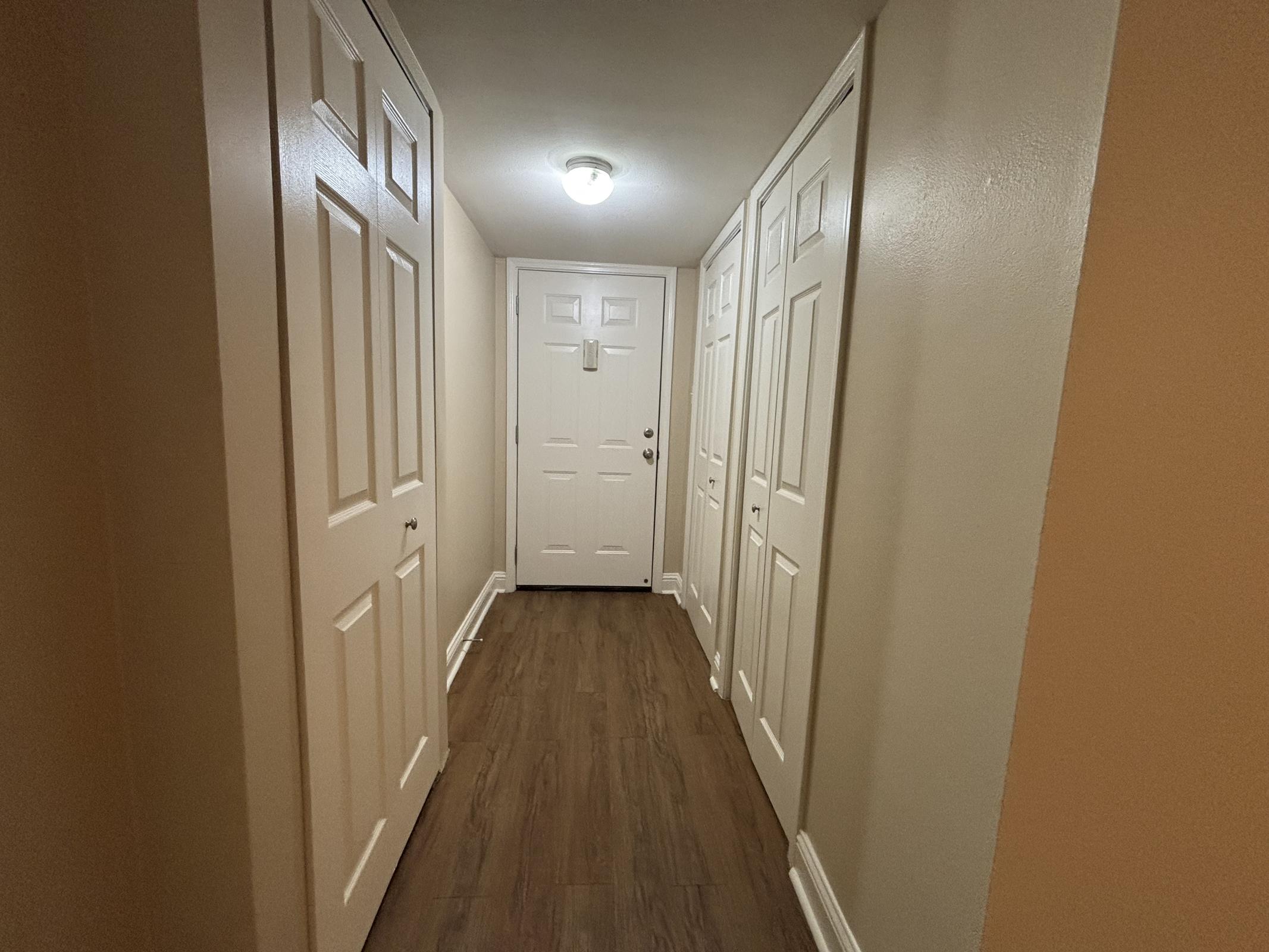 A narrow hallway featuring light-colored walls, wooden flooring, and multiple closet doors on one side. At the end of the hallway, there is a white door with a round light fixture above it, providing illumination. The space appears tidy and inviting.