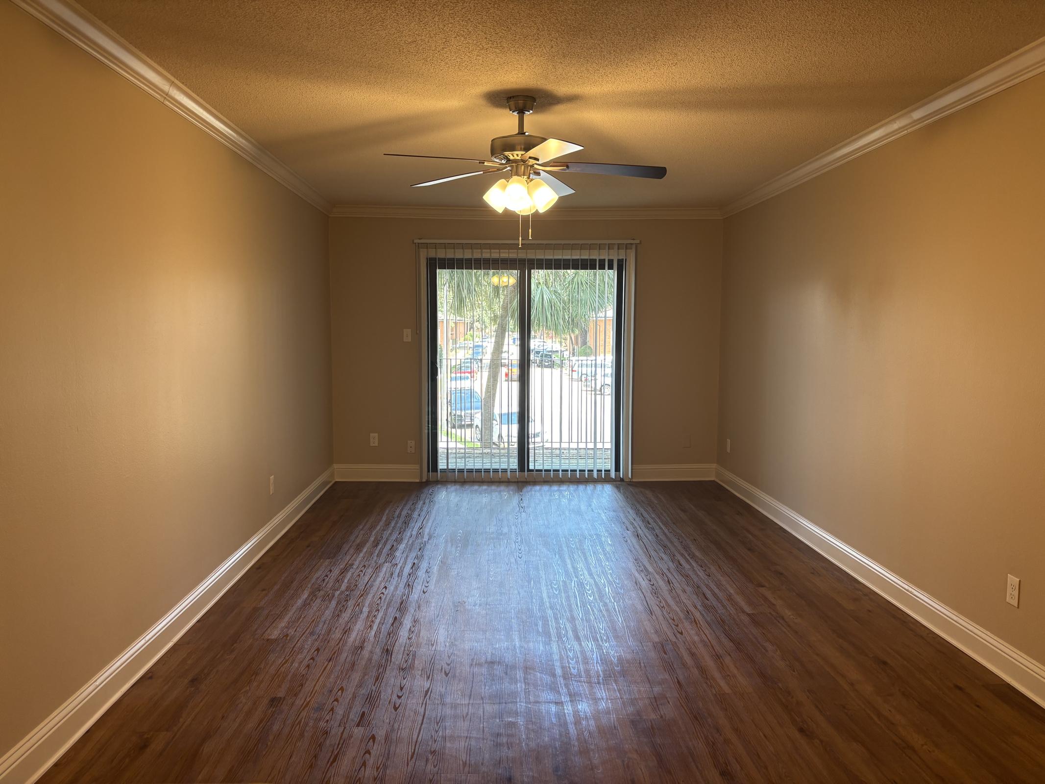 A spacious, empty living room with light brown walls and hardwood flooring. A ceiling fan with five light bulbs hangs from the ceiling. In the background, a sliding glass door leads to a patio, revealing a view of parked cars and greenery outside. The room is bright and well-lit.