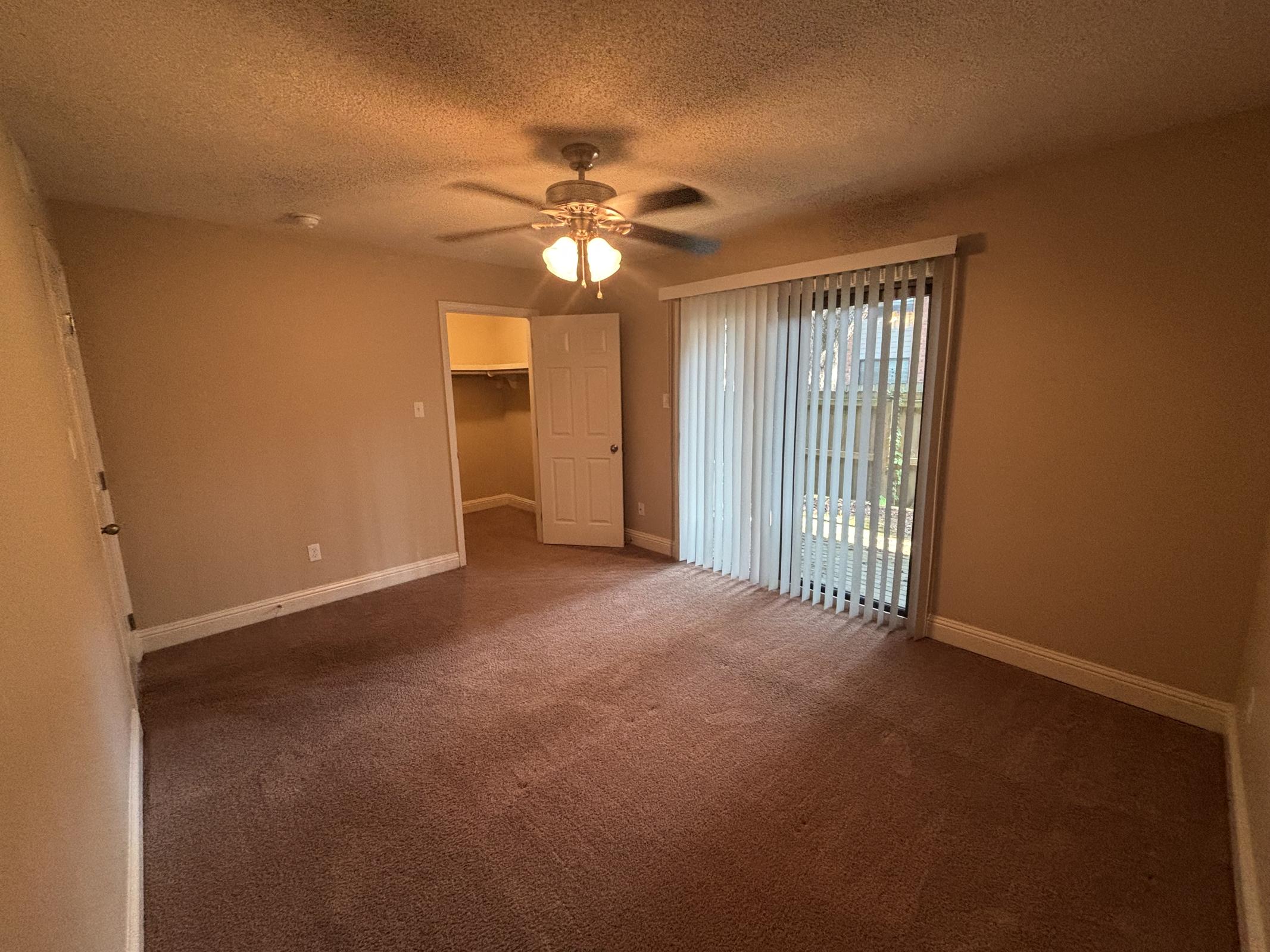 A spacious room with light brown walls and carpeting. There's a ceiling fan with light, a sliding glass door with vertical blinds leading to an outdoor area, and a closet door visible in the background. Natural light is coming in through the door, illuminating the room.